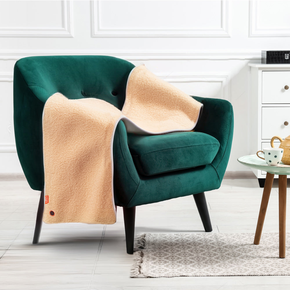 The COZY Heated Throw & Seat Cover Pad - JORV by Cozy Cushion EU is draped over a green velvet armchair in a bright, modern room with white walls, a small table with a polka-dot mug, dresser, and textured rug on the floor.