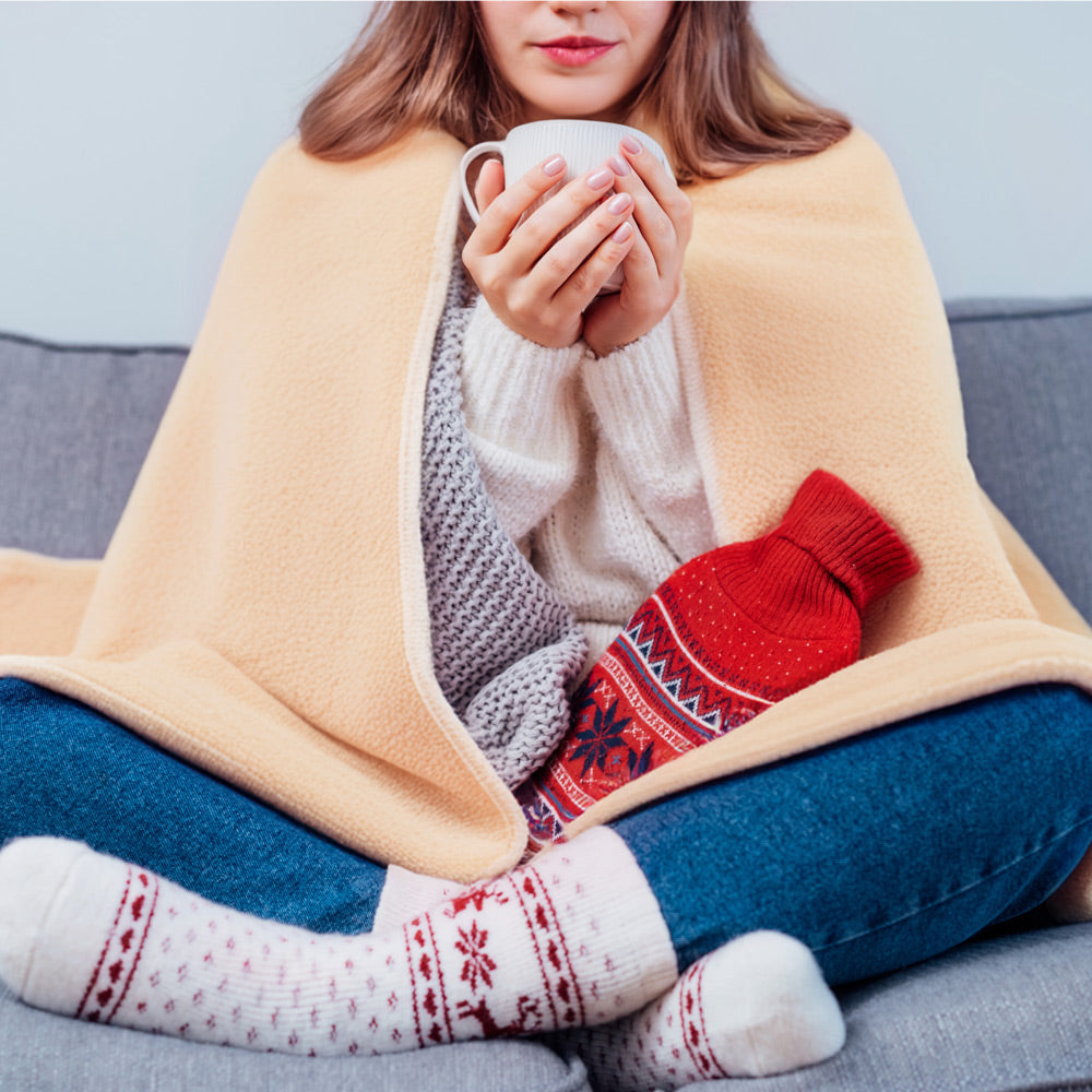 A woman sits cross-legged on a couch with the COZY Heated Throw & Seat Cover Pad by Cozy Cushion EU, holding a white mug. Dressed in winter clothes and patterned socks, she keeps warm in an eco-friendly way.