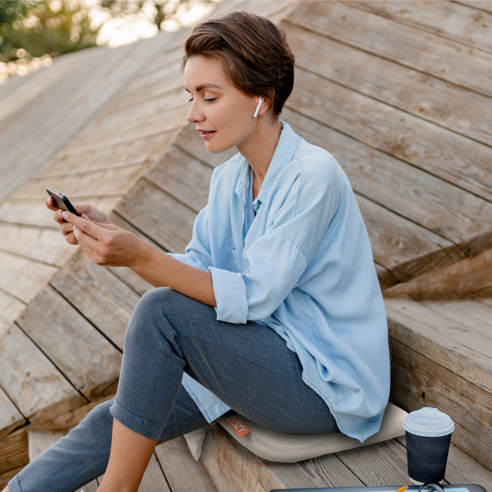 A woman with short hair, wearing wireless earbuds and a light blue shirt, sits on wooden steps, looking at her phone. Beside her is a takeaway coffee cup and the COZY Cordless Heated Seat Pad - JORD by Cozy Cushion EU for added comfort.