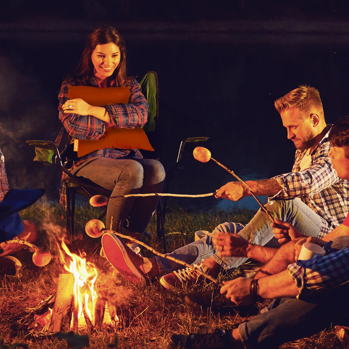 Four friends gather by a campfire at night, toasting marshmallows. One woman smiles, hugging the Cozy Cushion EU COZY Heated Cushion Cordless - UNA (45cm x 45cm), enjoying its rechargeable warmth while the others focus on roasting marshmallows.