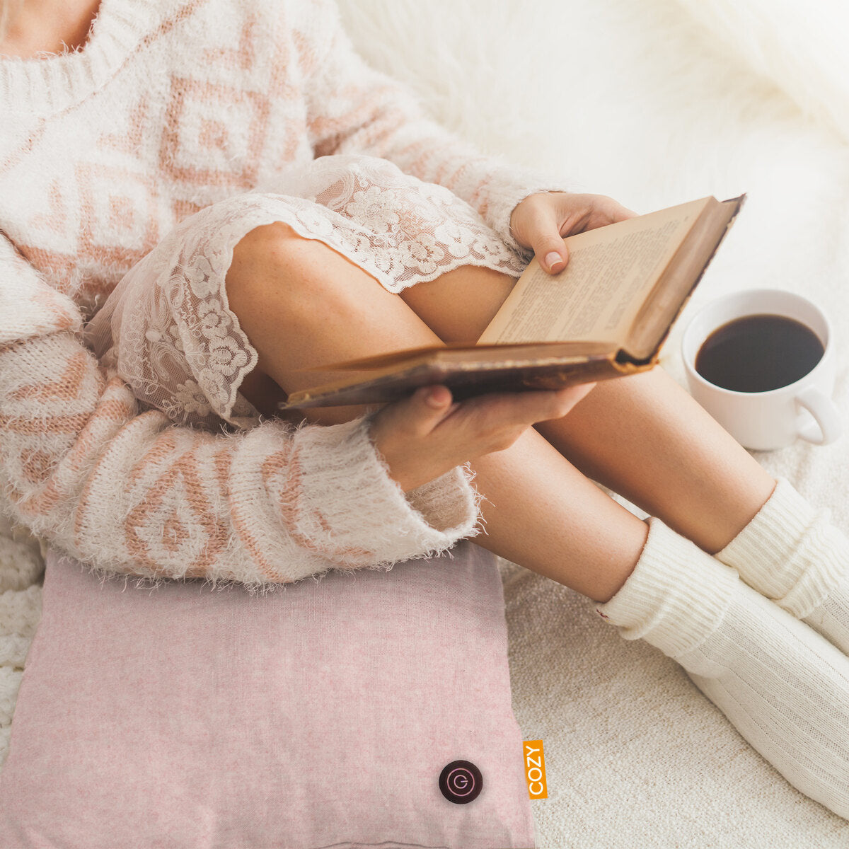 A person in a cozy sweater relaxes with a book and coffee, sitting on the COZY Heated Cushion Cordless - TOVE (60cm x 45cm) by Cozy Cushion EU. The scene feels warm, soft, and inviting.