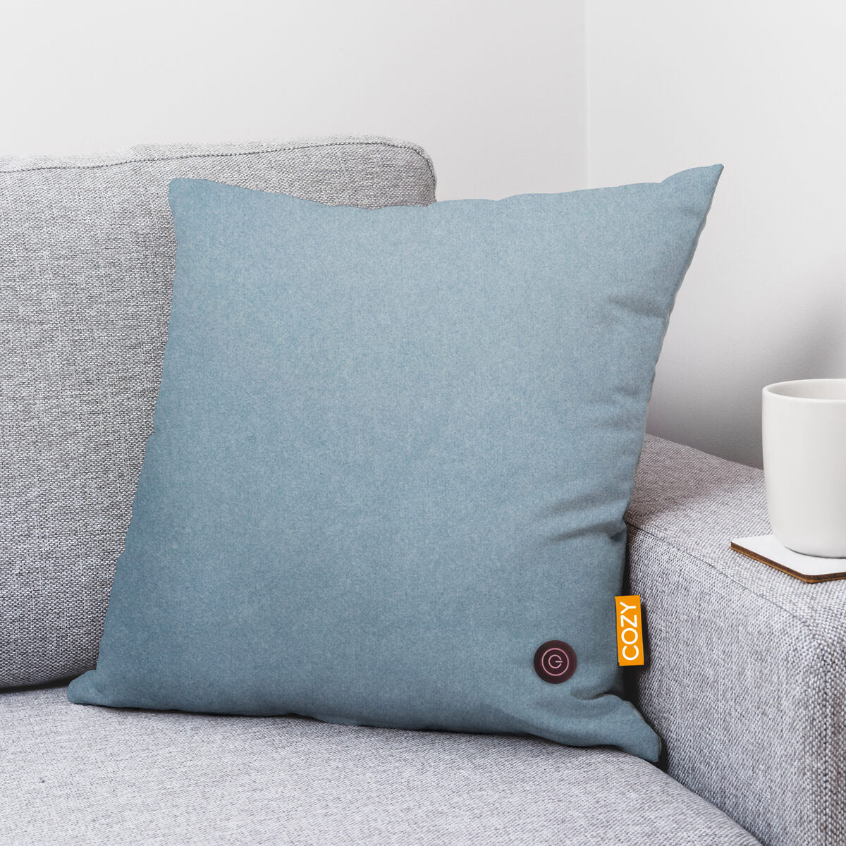 A light blue COZY Heated Cushion Cordless - UNA (45cm x 45cm) by Cozy Cushion EU rests on a light gray sofa, with a white mug on a coaster placed on the armrest near the cushion.