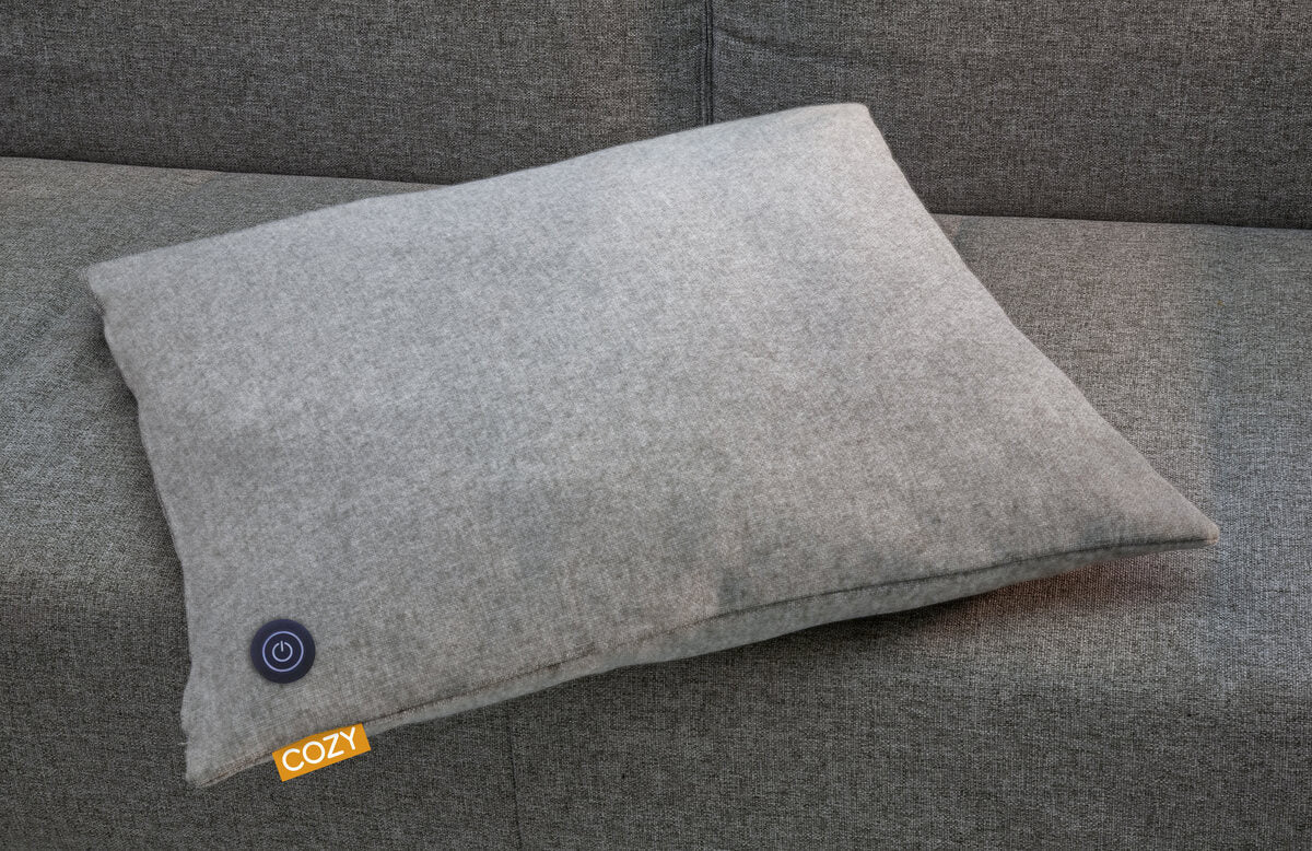 A gray COZY Heated Cushion Cordless - TOVE (60cm x 45cm) from Cozy Cushion rests on a matching sofa. It features a circular power button symbol and a yellow COZY tag in the corner.