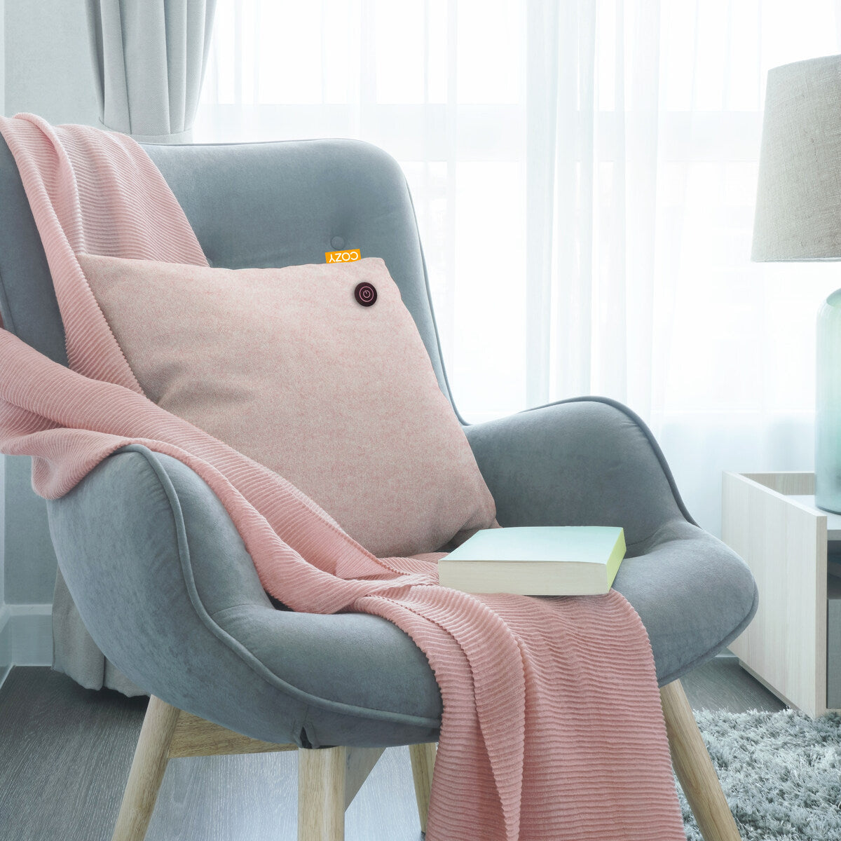A cozy gray armchair with wooden legs holds a pink pillow and the COZY Heated Cushion Cordless - UNA Velvet (45cm x 45cm) by Cozy Cushion EU. A closed book rests on the seat, framed by a lamp and window with sheer curtains.