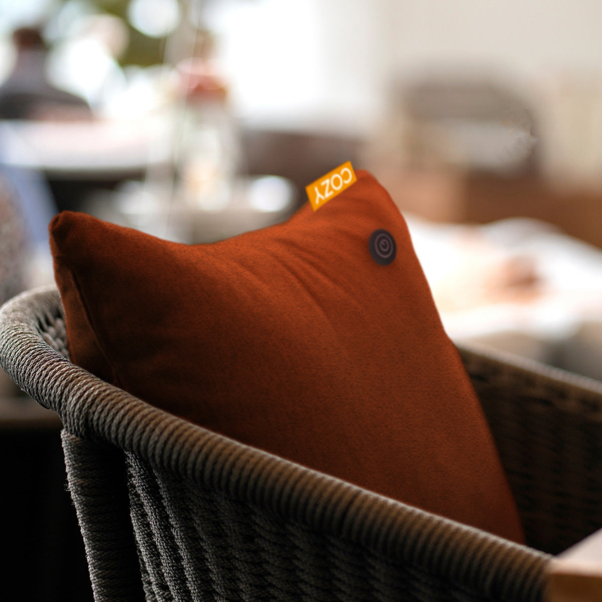 The Cozy Cushion COZY Heated Cushion Cordless - TOVE (60cm x 45cm) by Cozy Cushion, in brown with a small black button and yellow COZY tag, provides gentle infrared warmth on a woven chair in a softly lit indoor space.