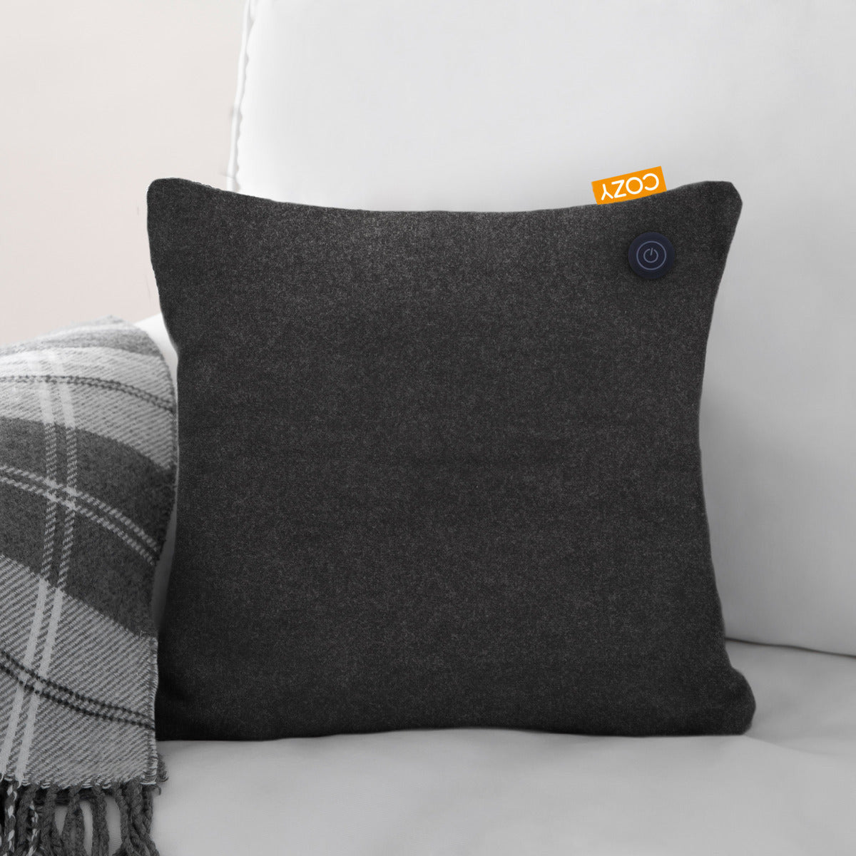 The COZY Heated Cushion Cordless - UNA (45cm x 45cm) by Cozy Cushion EU, in dark gray with an orange COZY tag, offers cordless heated comfort and a rechargeable battery, resting on a couch beside a plaid blanket.