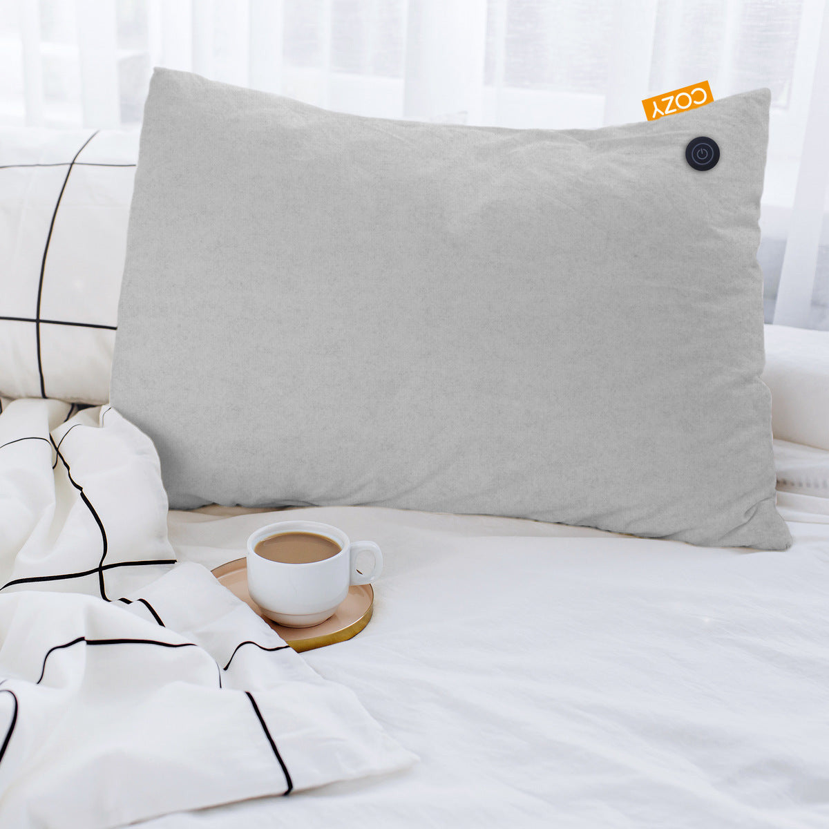 A COZY Heated Cushion Cordless - TOVE (60cm x 45cm) by Cozy Cushion EU with a round black button rests on white plaid bedding beside a coffee cup on a coaster, with white curtains in the background.
