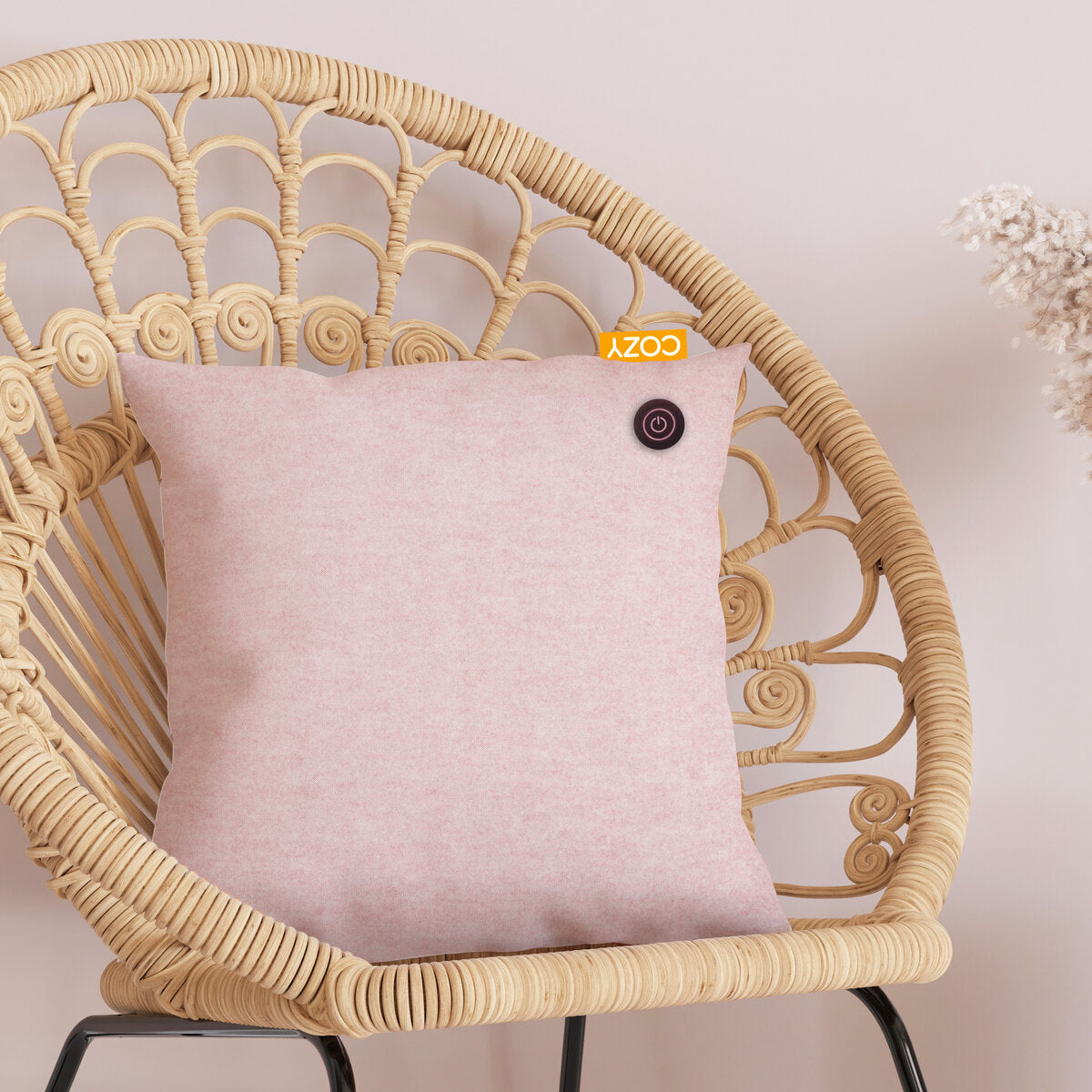 The COZY Heated Cushion Cordless - UNA Velvet (45cm x 45cm) by Cozy Cushion EU, in light pink with a small round button and yellow COZY tag, sits on a rattan chair against a pale pink wall beside dried flowers.