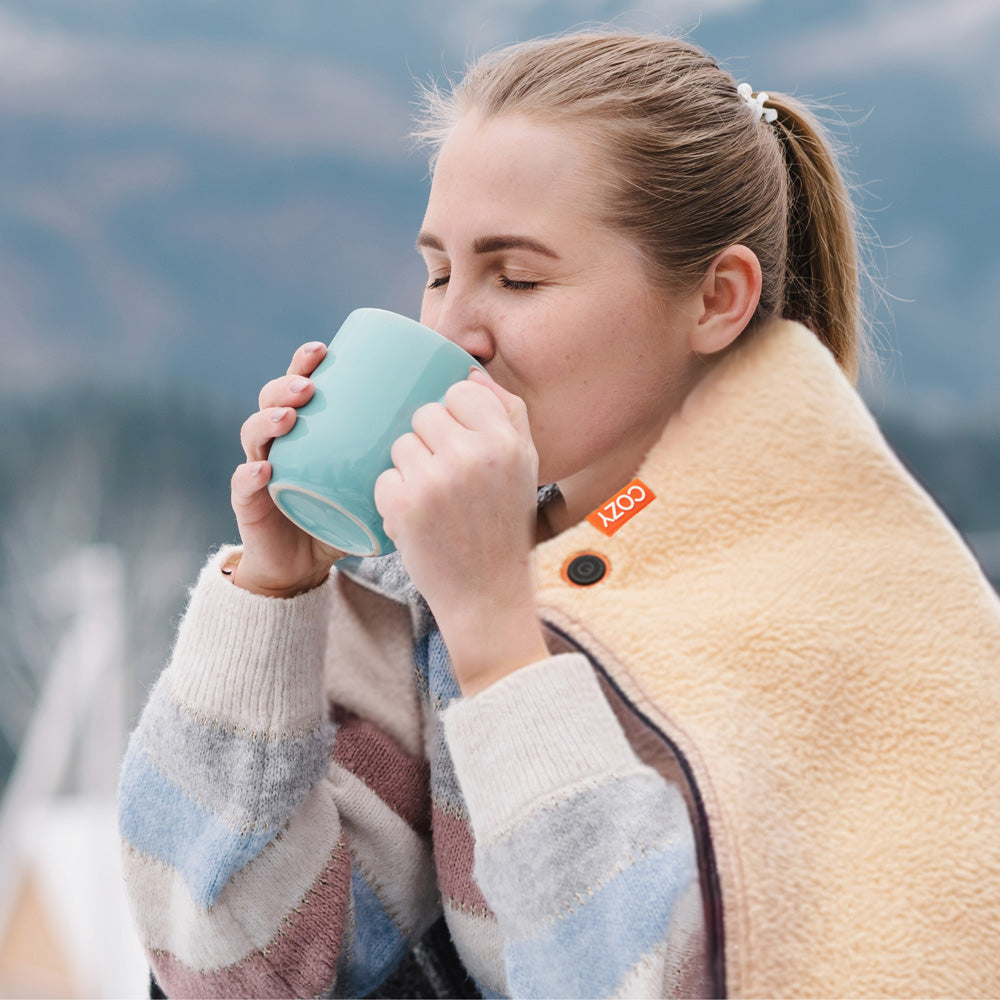 A woman wrapped in the COZY Heated Throw & Seat Cover Pad by Cozy Cushion EU enjoys a warm drink from a light blue mug outdoors, wearing a striped sweater with snowy mountains blurred in the background.