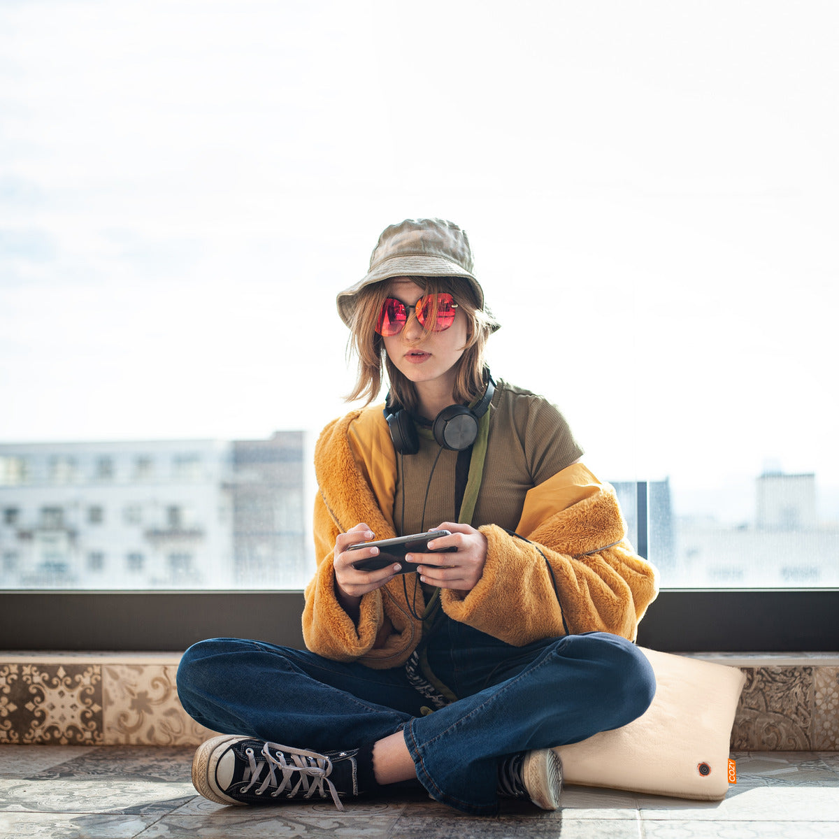 A young person in a yellow jacket, bucket hat, red sunglasses, headphones, jeans, and sneakers sits cross-legged by a window on the COZY Heated Cushion Cordless - UNA Faux Fur (45cm x 45cm) by Cozy Cushion EU, playing a handheld game console.