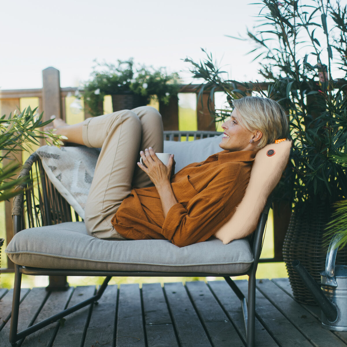 A woman relaxes on a wooden deck surrounded by greenery, smiling with her legs up and a mug in hand, leaning against the COZY Heated Cushion Cordless - UNA Faux Fur (45cm x 45cm) from Cozy Cushion EU.