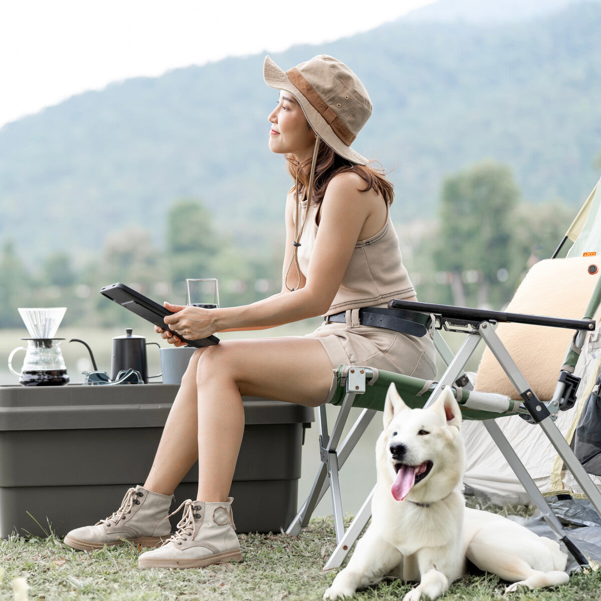 A woman in beige outdoor gear sits on a camping chair with a tablet and coffee, using the COZY Heated Cushion Cordless - UNA Faux Fur (45cm x 45cm) by Cozy Cushion EU. A white dog relaxes on the grass, surrounded by trees and mountains.