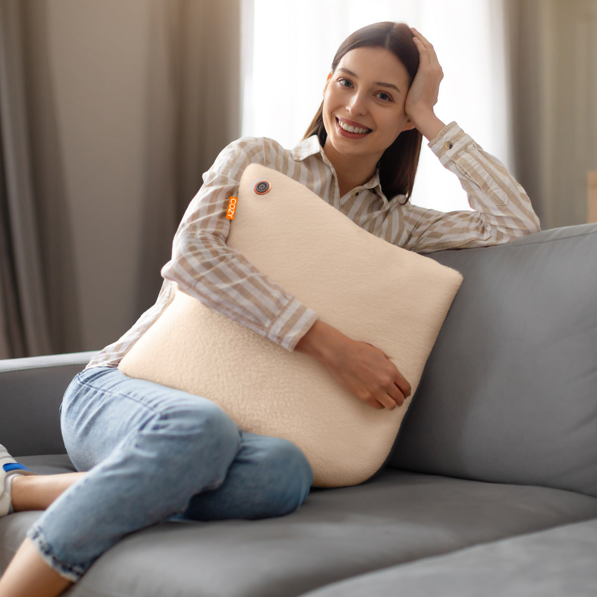 A woman in a striped shirt and jeans smiles while hugging the Cozy Cushion EU COZY Heated Cushion Cordless - UNA Faux Fur (45cm x 45cm) in beige, relaxing comfortably on her couch in a cozy, well-lit living room.