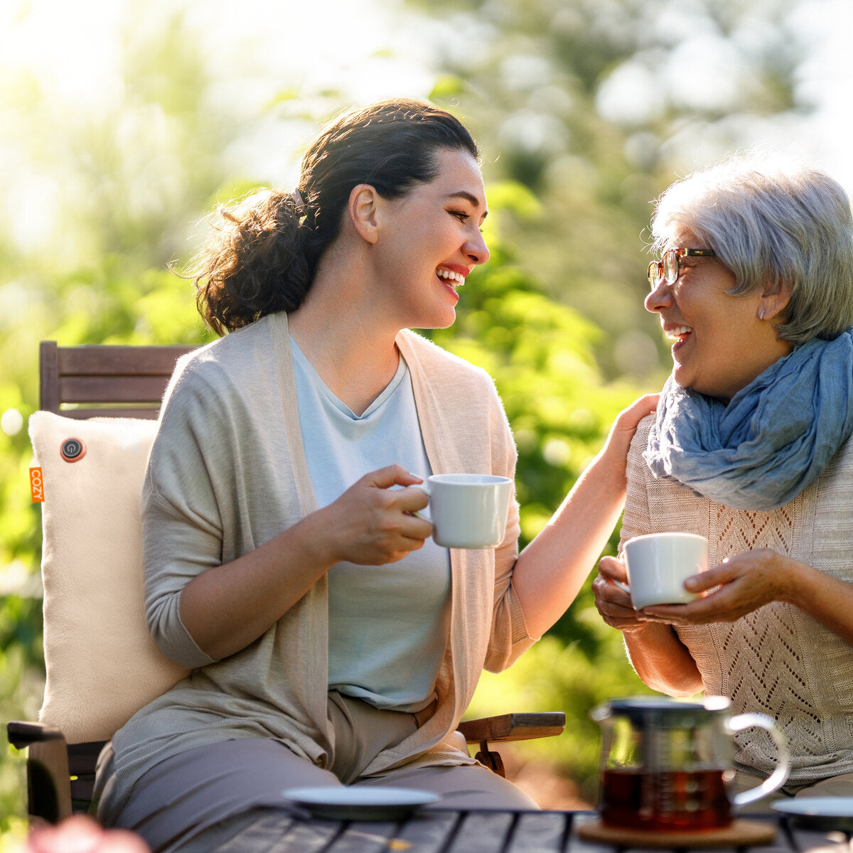 Two women enjoy tea in a sunny garden, smiling on wooden chairs. The older woman relaxes with the COZY Heated Cushion Cordless - UNA Faux Fur (45cm x 45cm) from Cozy Cushion EU, its infrared warmth enhancing the cheerful atmosphere.
