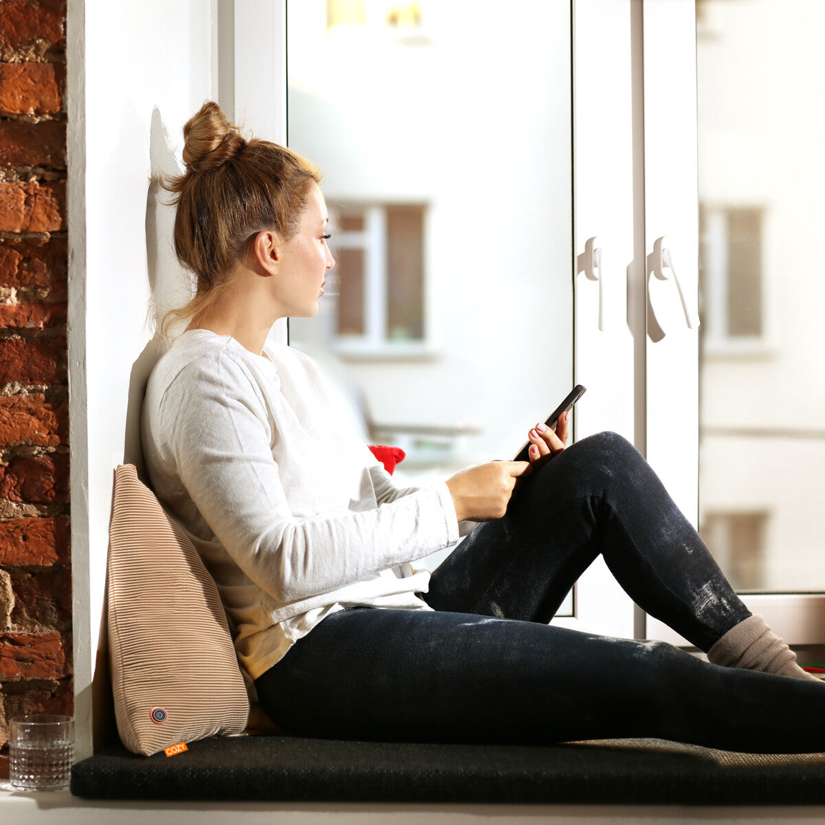 A blonde woman with her hair in a bun relaxes on a window seat, leaning against a pillow and using her smartphone. She sits on the COZY Heated Cushion Cordless - UNA Knitted (45cm x 45cm) by Cozy Cushion EU. A glass of water is beside her.