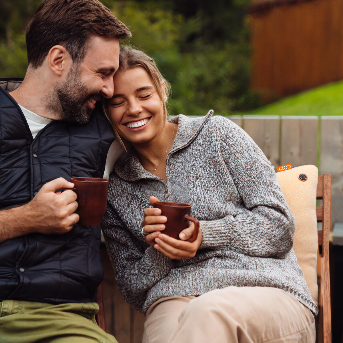 A smiling couple enjoys a cozy moment outdoors, holding mugs and leaning together on the COZY Heated Cushion Cordless - UNA (45cm x 45cm) by Cozy Cushion EU, powered by a rechargeable battery. The woman wears a gray sweater; the man wears a dark vest.