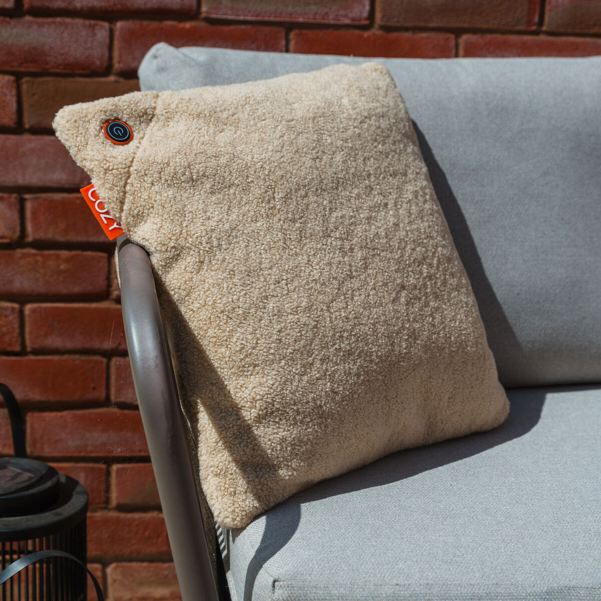 The COZY Heated Cushion Cordless - UNA Woolly (45cm x 45cm) by Cozy Cushion EU, a beige textured pillow with an orange tag and circular logo, rests on a light gray outdoor chair. A brick wall and black lantern are visible in the background.