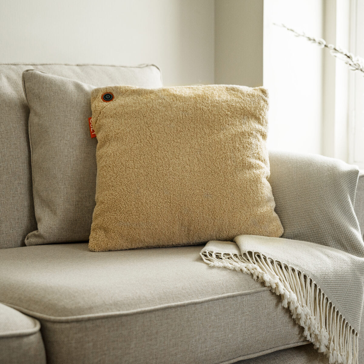 A beige textured pillow with button detail sits on a light gray sofa beside the Cozy Cushion EU COZY Heated Cushion Cordless - UNA Woolly (45cm x 45cm) and a cream fringed throw, in a softly lit living room.