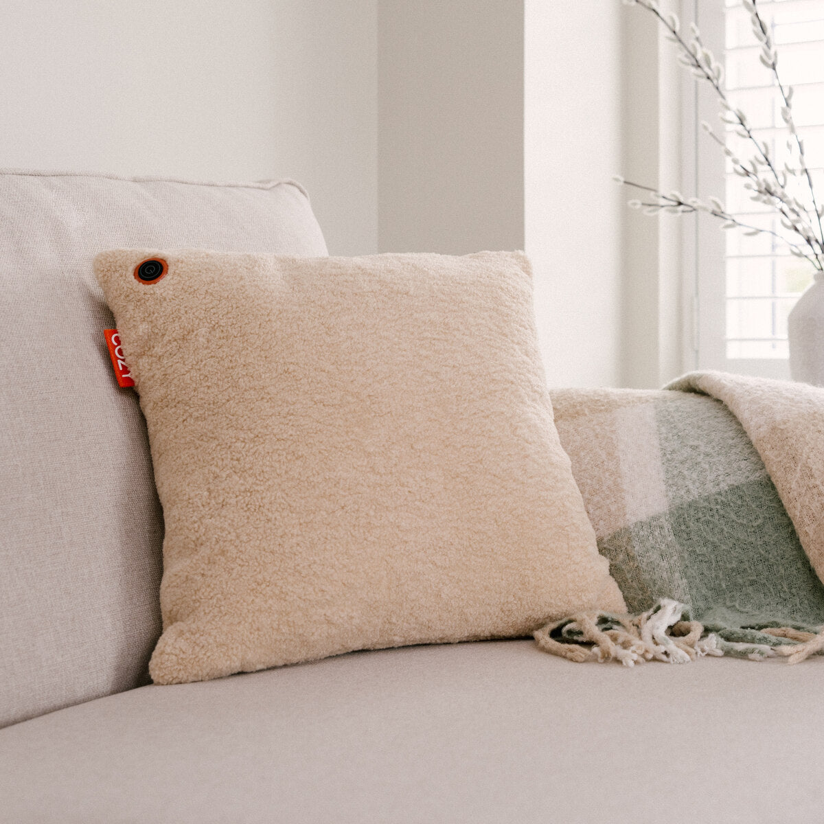 A beige, textured square pillow with a red tag sits on a light sofa. Nearby, a green-and-white plaid blanket and the COZY Heated Cushion Cordless - UNA Woolly (45cm x 45cm) by Cozy Cushion EU enjoy the soft natural window light.