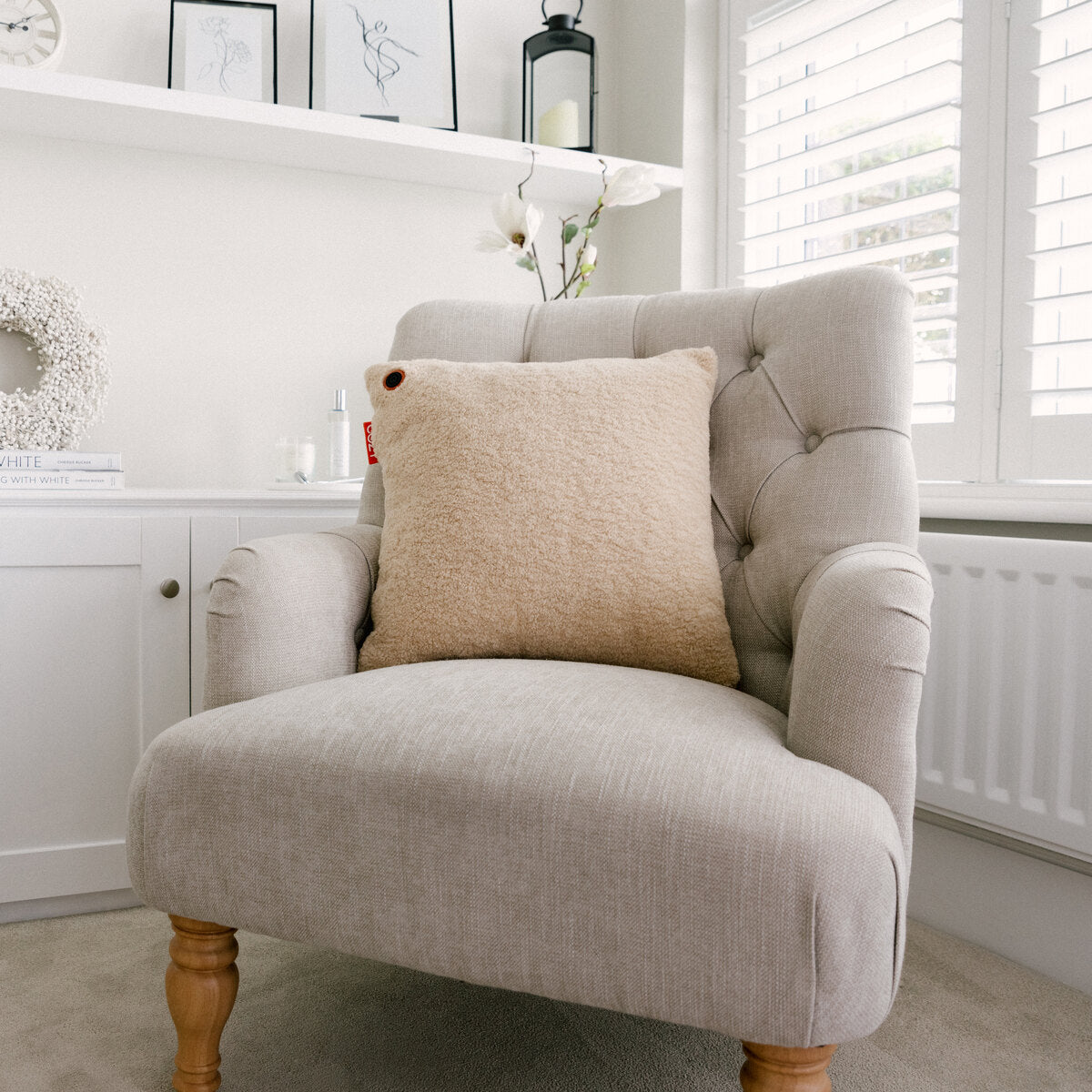 A beige, tufted armchair with wooden legs sits by a window, topped with the COZY Heated Cushion Cordless - UNA Woolly (45cm x 45cm) by Cozy Cushion EU. Shelves behind display books, framed art, and minimalist decor.