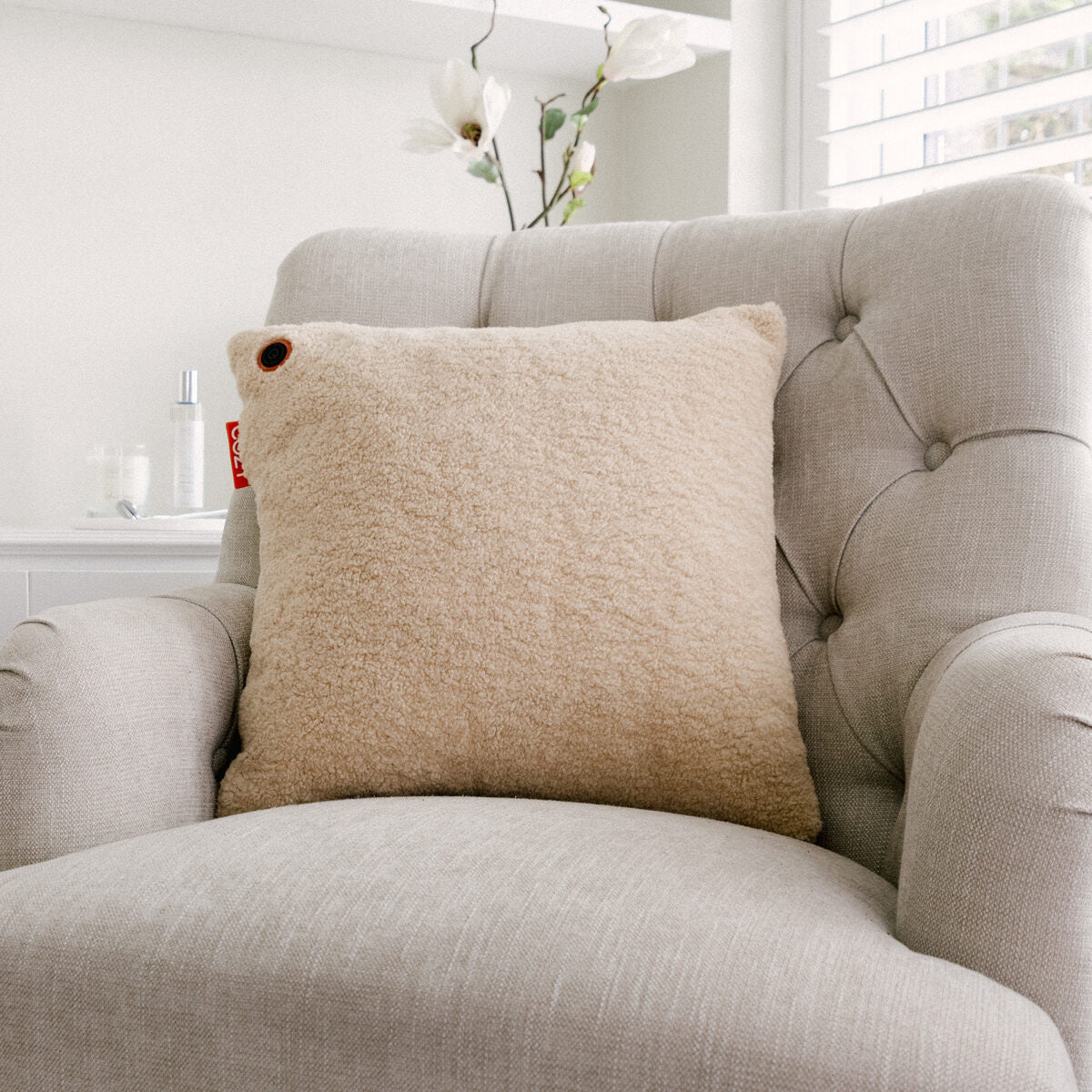 The COZY Heated Cushion Cordless - UNA Woolly (45cm x 45cm) by Cozy Cushion EU, in beige textured fabric with a rechargeable battery, is displayed on a light gray tufted armchair in a bright room with white flowers and window blinds.