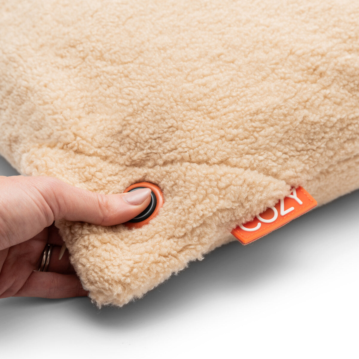 A hand presses a black button on the corner of the Cozy Cushion EU COZY Heated Cushion Cordless - UNA Woolly (45cm x 45cm), a soft, beige fleece-like pillow with an orange COZY tag, offering rechargeable, portable warmth anywhere.