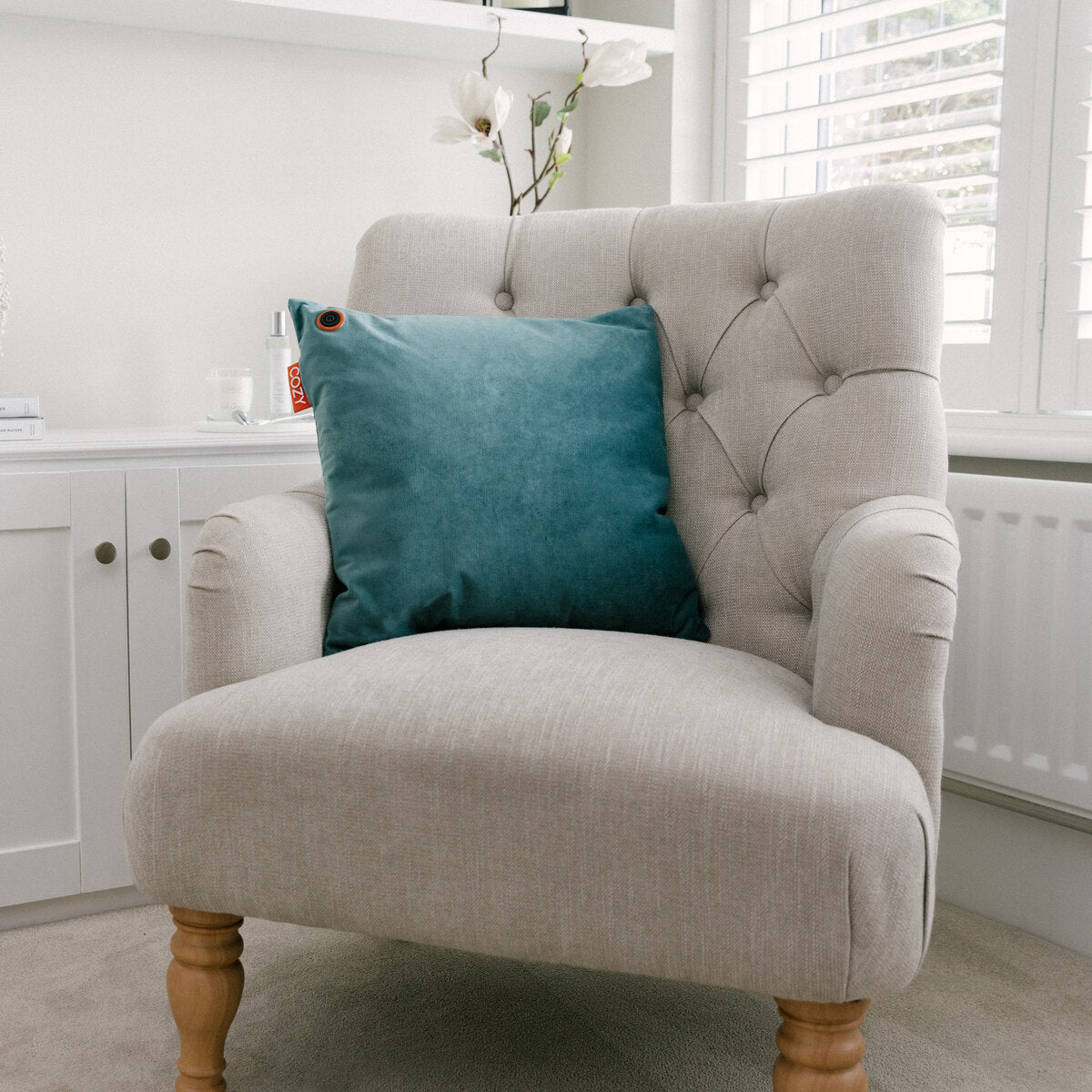A beige upholstered armchair with wooden legs features the COZY Heated Cushion Cordless - UNA Velvet (45cm x 45cm) by Cozy Cushion EU. Its placed near a window with white shutters, a white cabinet, and an orchid in the background.