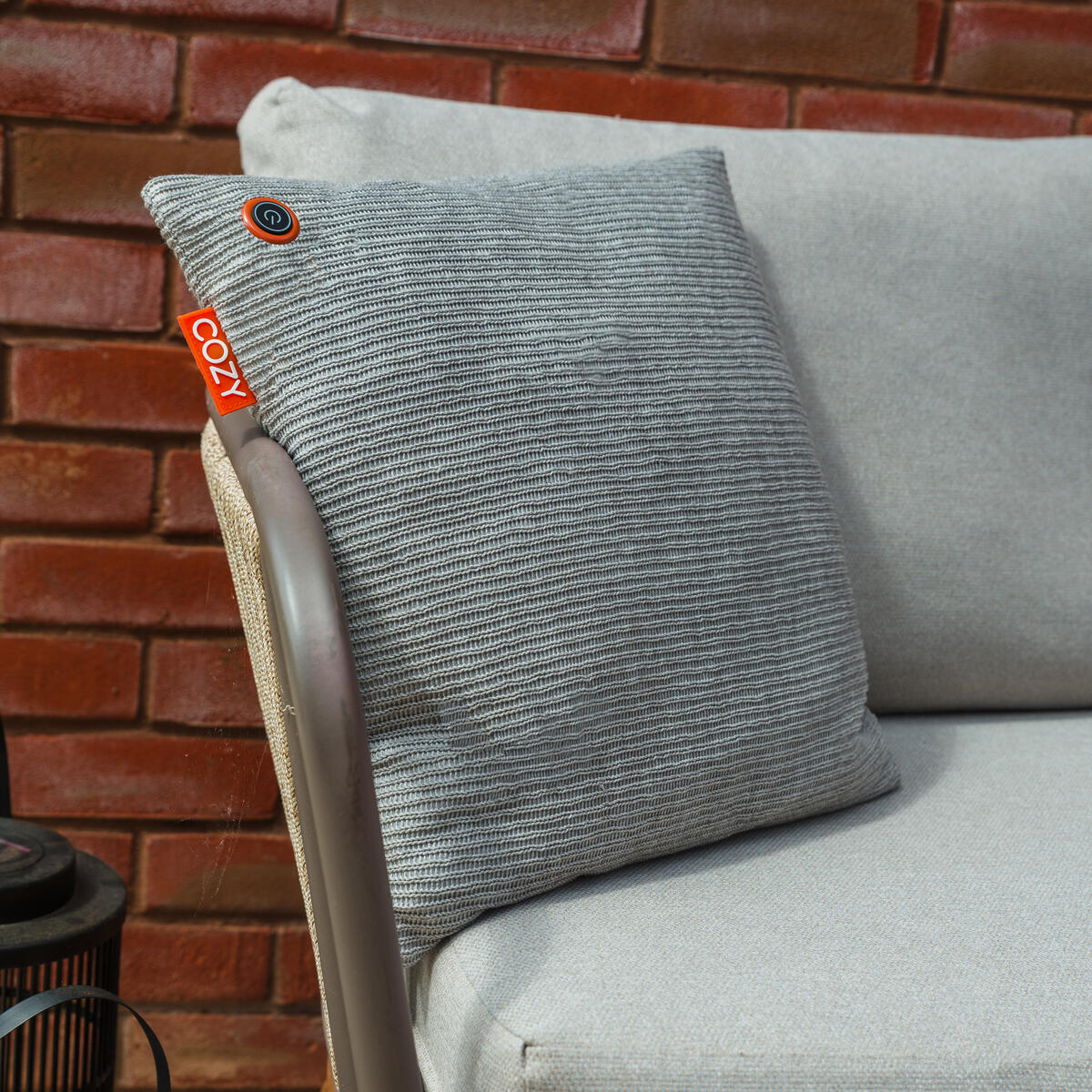The COZY Heated Cushion Cordless - UNA Knitted (45cm x 45cm) by Cozy Cushion EU, featuring a gray textured cover and orange COZY tag, sits on a light gray outdoor chair before a red brick wall.