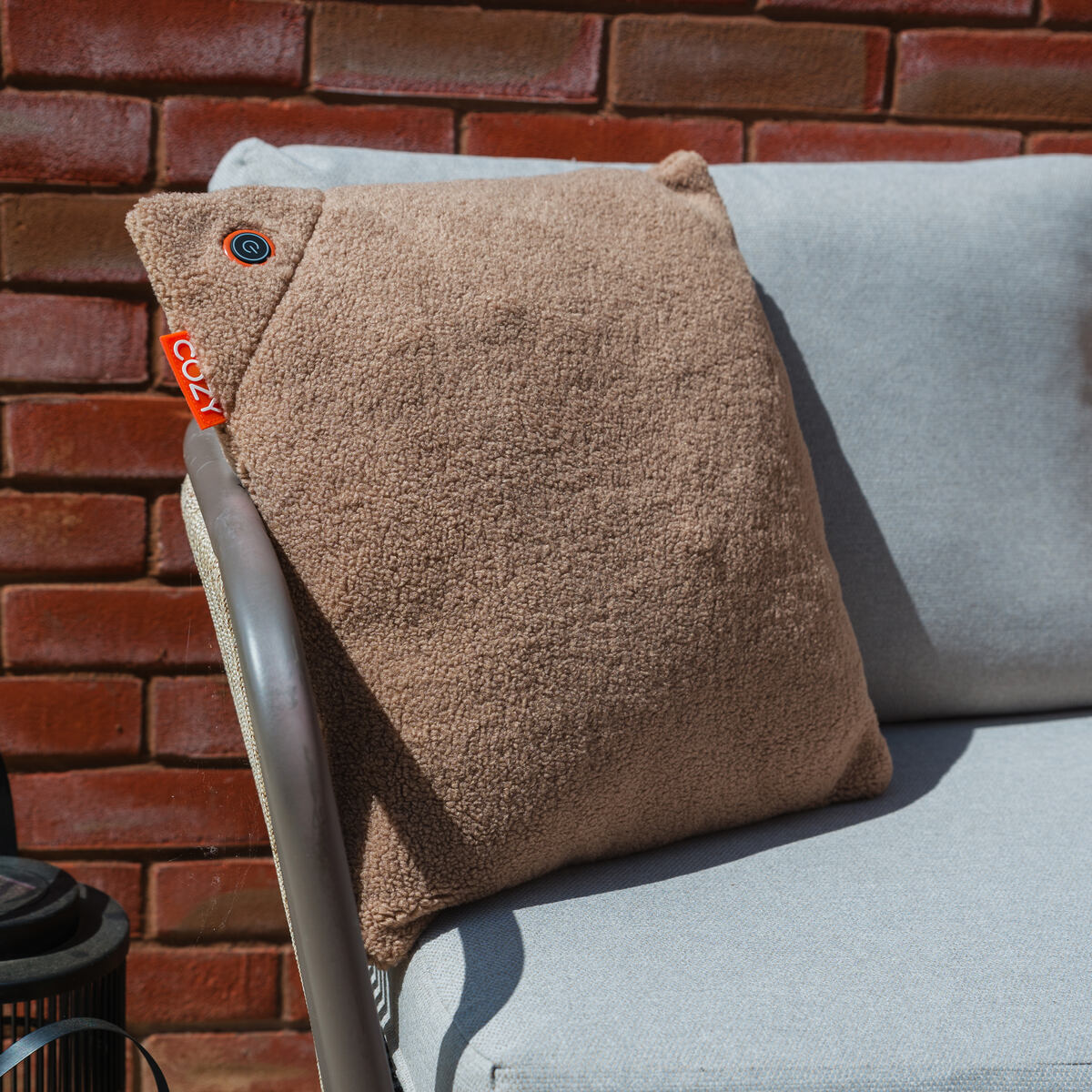 A COZY Heated Cushion Cordless - UNA Woolly (45cm x 45cm) by Cozy Cushion EU in soft brown rests on a light gray outdoor chair, set against a sunlit red brick wall.