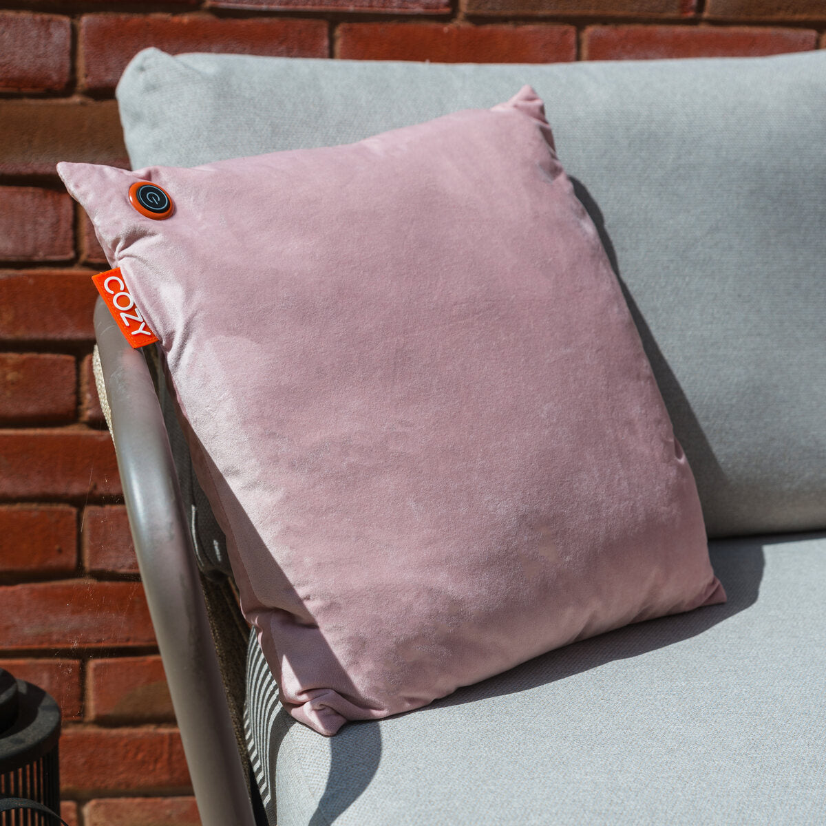 The COZY Heated Cushion Cordless - UNA Velvet (45cm x 45cm) by Cozy Cushion EU, in light pink with a COZI tag and round button, sits on a light gray outdoor sofa against a red brick wall.