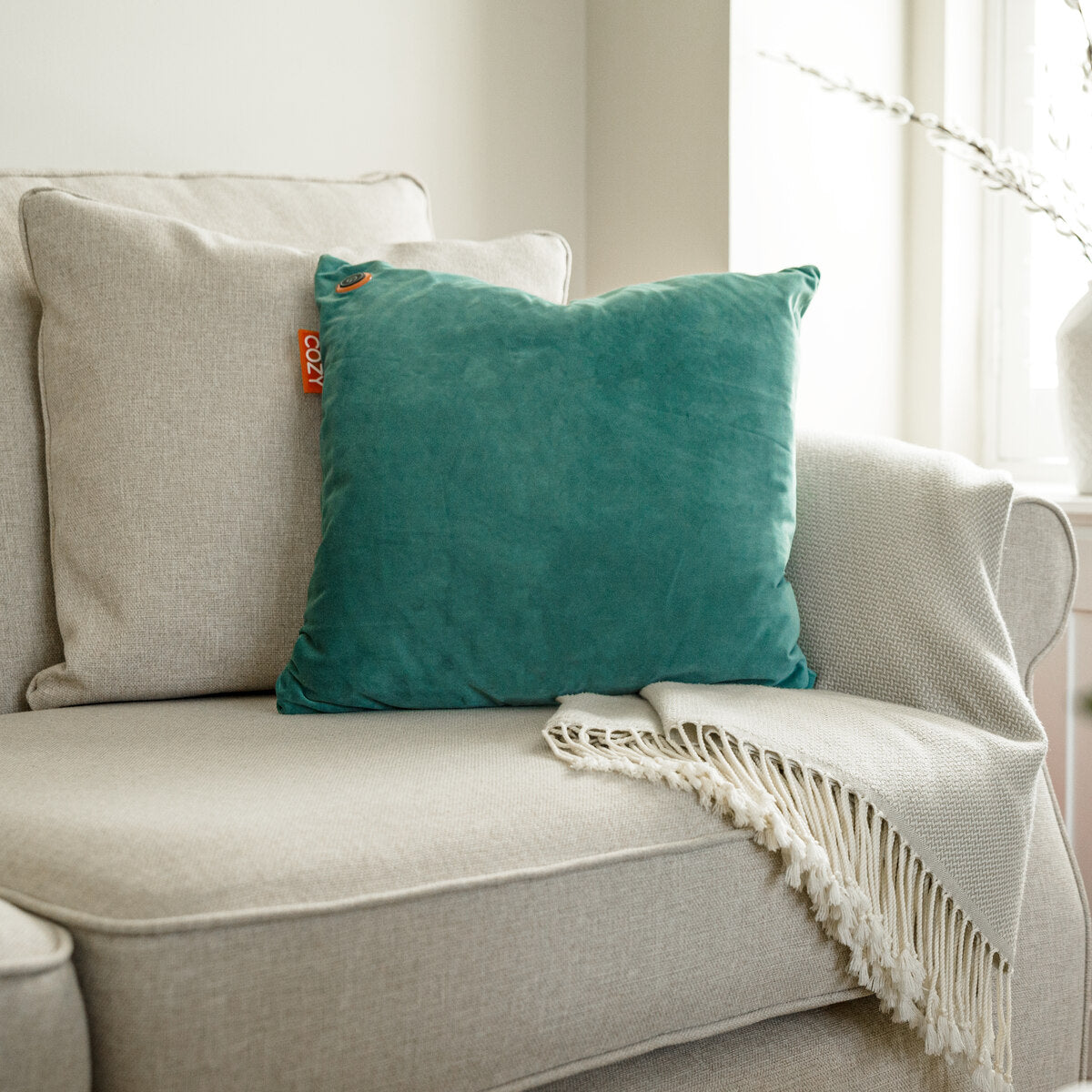 The COZY Heated Cushion Cordless - UNA Velvet (45cm x 45cm) by Cozy Cushion EU in teal, featuring a small orange tag, rests on a light beige sofa with a textured cream blanket near a sunlit window.