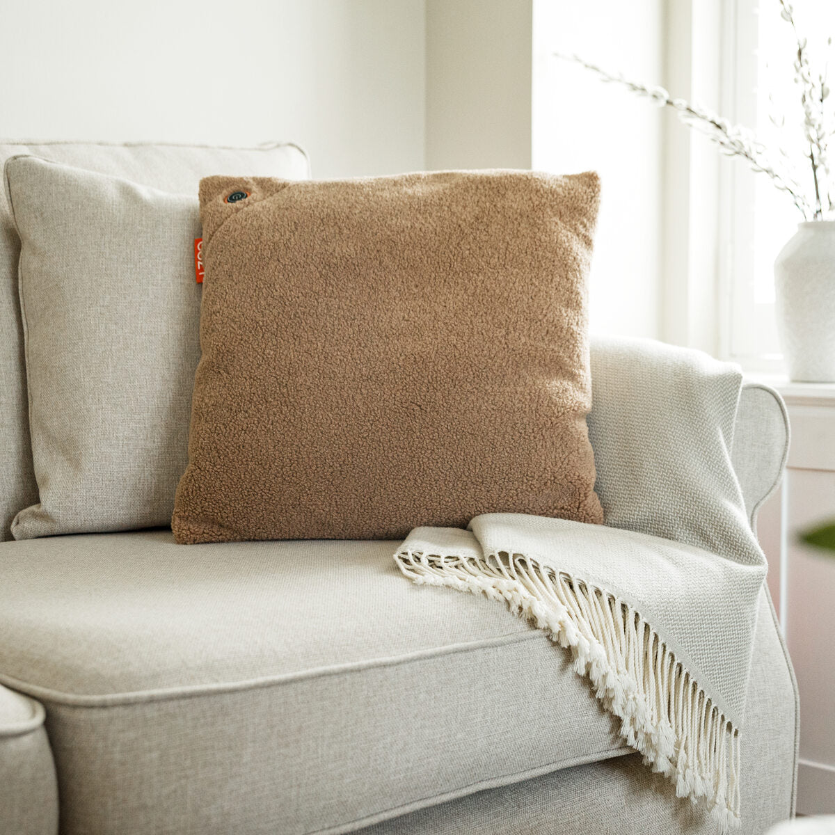 A beige armchair features the COZY Heated Cushion Cordless - UNA Woolly (45cm x 45cm) by Cozy Cushion EU in light brown, paired with a cream fringed throw, set near a sunlit window and a white vase in the background.