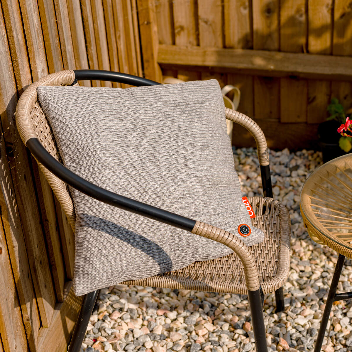 A wicker chair with black metal arms holds the COZY Heated Cushion Cordless - UNA Knitted (45cm x 45cm) by Cozy Cushion EU in light gray. The chair sits outdoors on gravel beside a small round wicker table, with a wooden fence behind.