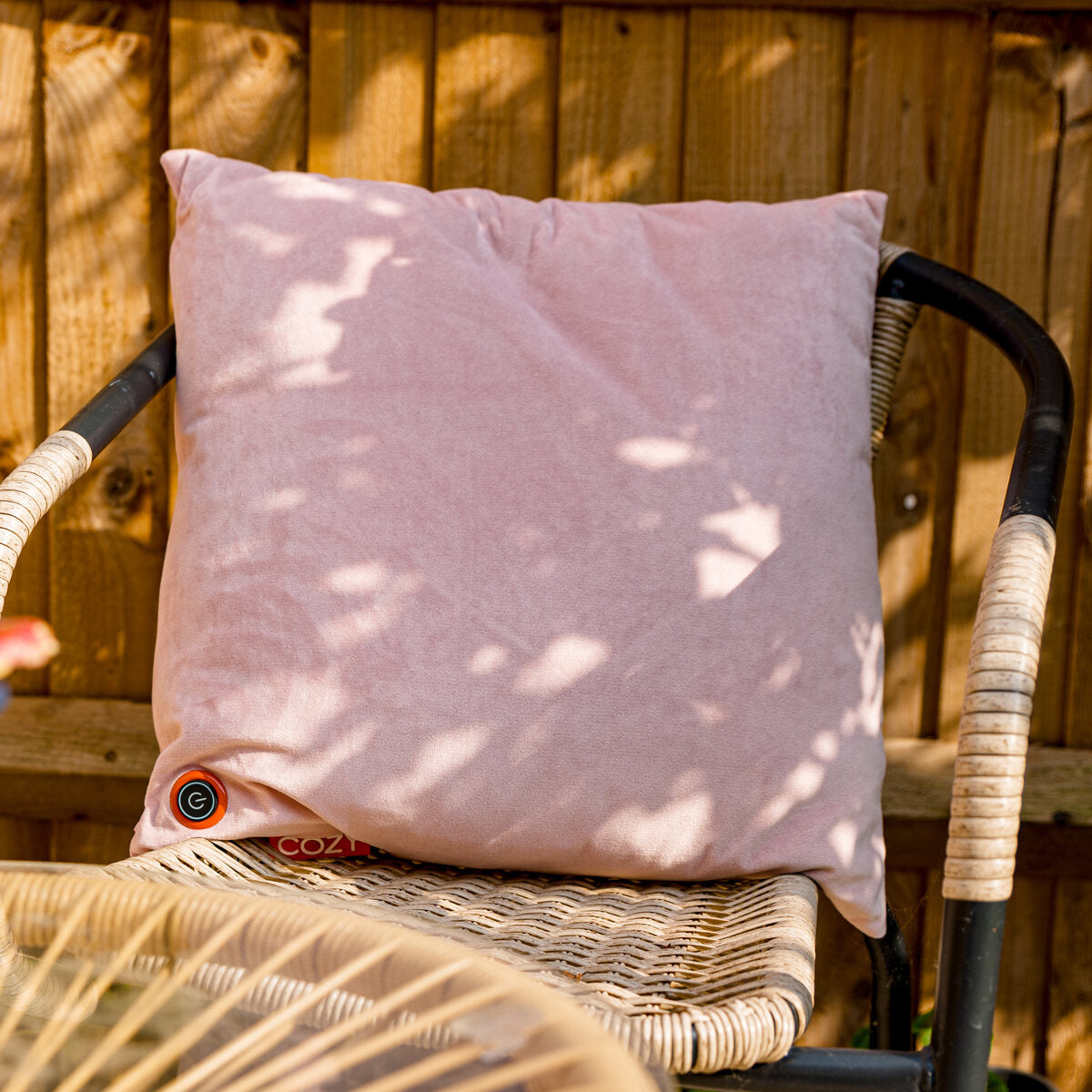 The COZY Heated Cushion Cordless - UNA Velvet (45cm x 45cm) by Cozy Cushion EU in light pink sits on a wicker chair with black armrests outdoors, sunlight casting soft shadows across the cushion and chair.