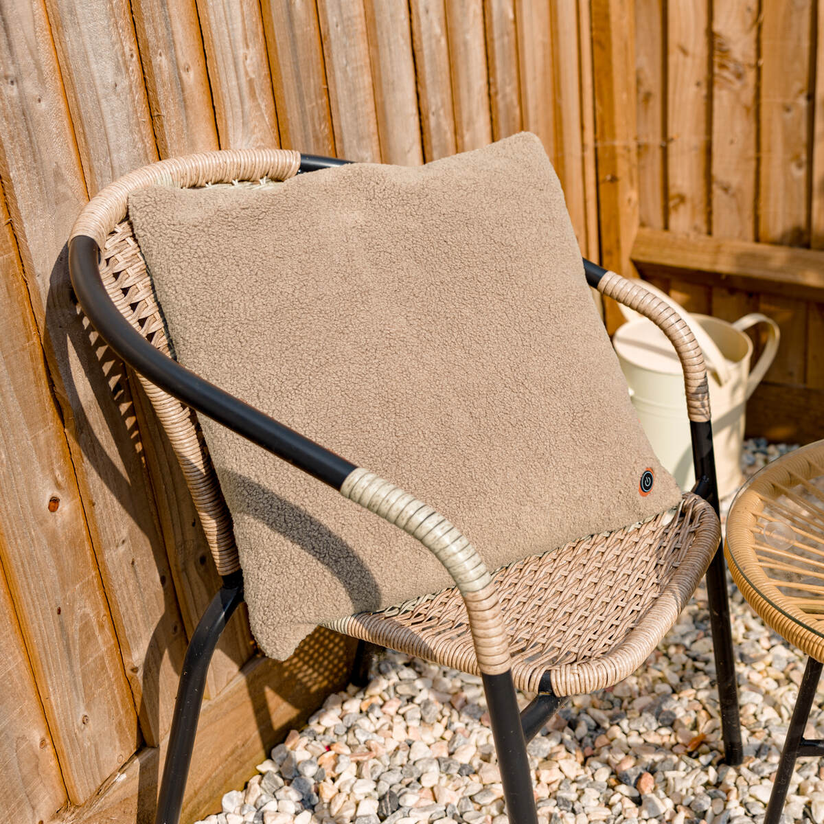 The COZY Heated Cushion Cordless - UNA Woolly (45cm x 45cm) by Cozy Cushion EU, in beige, is shown on a black-framed wicker chair beside a wooden fence and white watering can on a gravel surface.