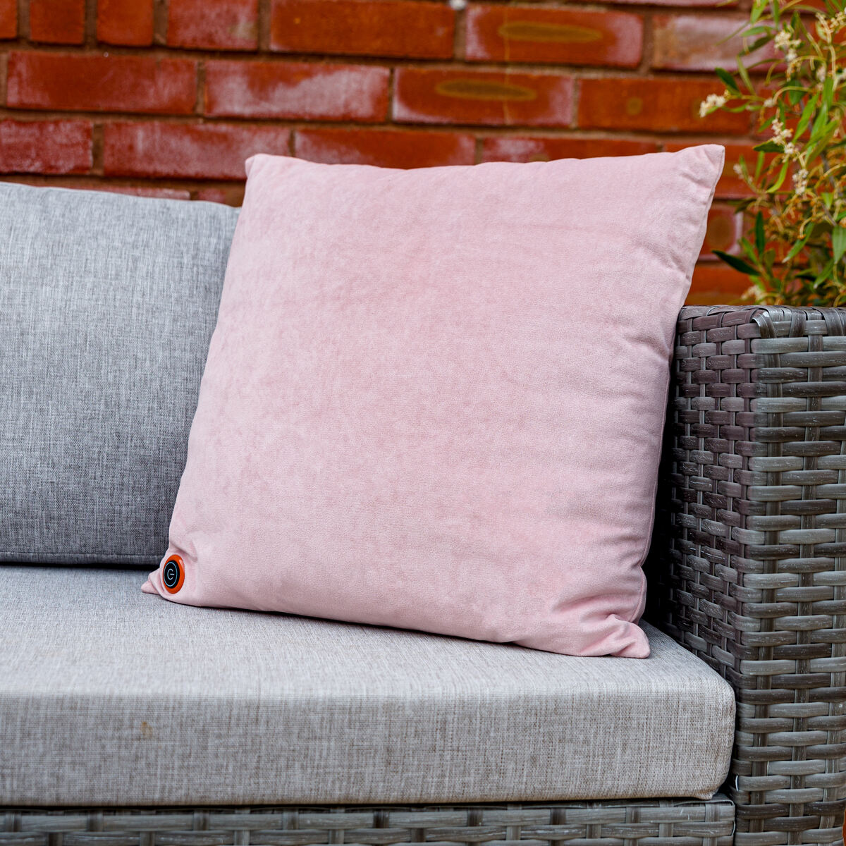 The COZY Heated Cushion Cordless - UNA Velvet (45cm x 45cm) by Cozy Cushion EU in light pink sits on a gray wicker outdoor sofa, showing a small round patch. A red brick wall and green plants appear in the background.
