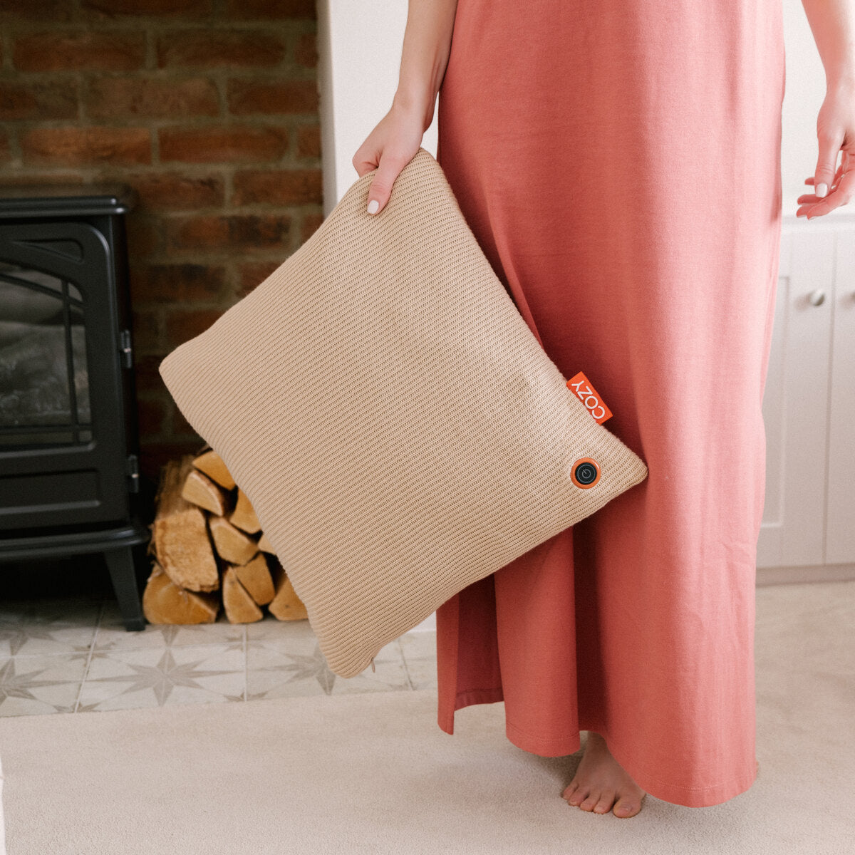 A person in a pink dress stands barefoot indoors, holding the Cozy Cushion EU COZY Heated Cushion Cordless - UNA Knitted (45cm x 45cm); firewood and a black stove are in the background.