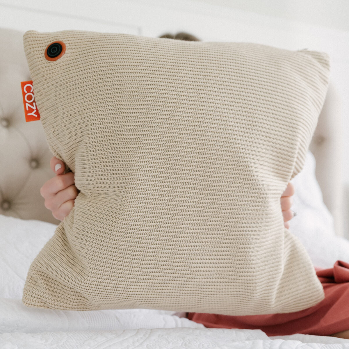 A person sits on a bed holding the Cozy Cushion EU COZY Heated Cushion Cordless - UNA Knitted (45cm x 45cm), a plush beige pillow with infrared technology and rechargeable battery for ultimate comfort.