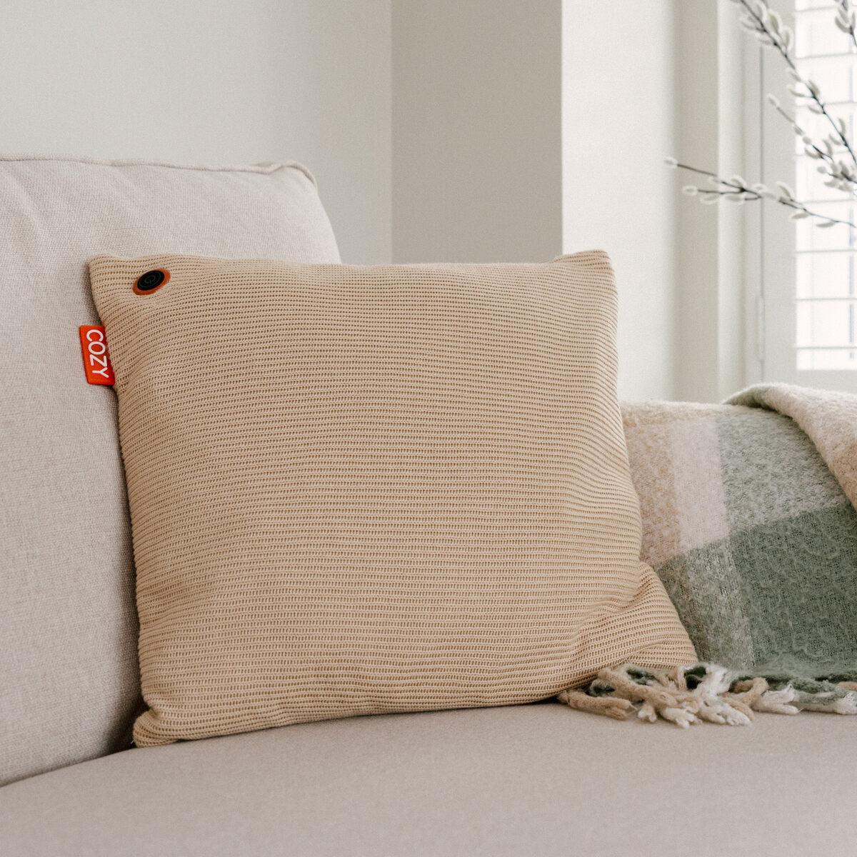 The Cozy Cushion EU COZY Heated Cushion Cordless - UNA Knitted (45cm x 45cm), featuring a beige textured cover and red COZY tag, is displayed on a light sofa beside a green and white plaid blanket, with a window and plant behind.