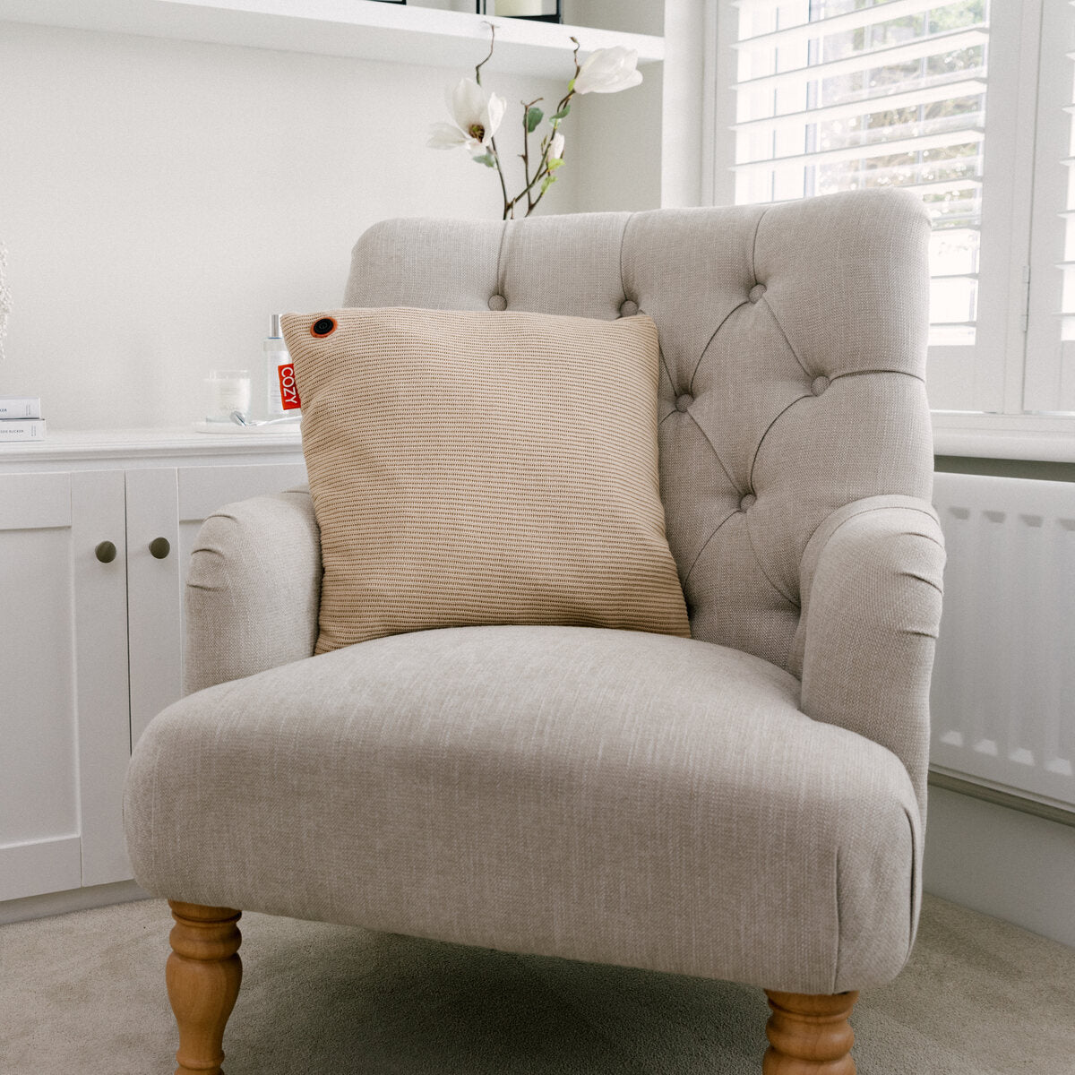 A light beige armchair with wooden legs sits in a bright room, featuring the COZY Heated Cushion Cordless - UNA Knitted (45cm x 45cm) by Cozy Cushion EU. In the background are white cabinetry, a potted orchid, and window shutters.