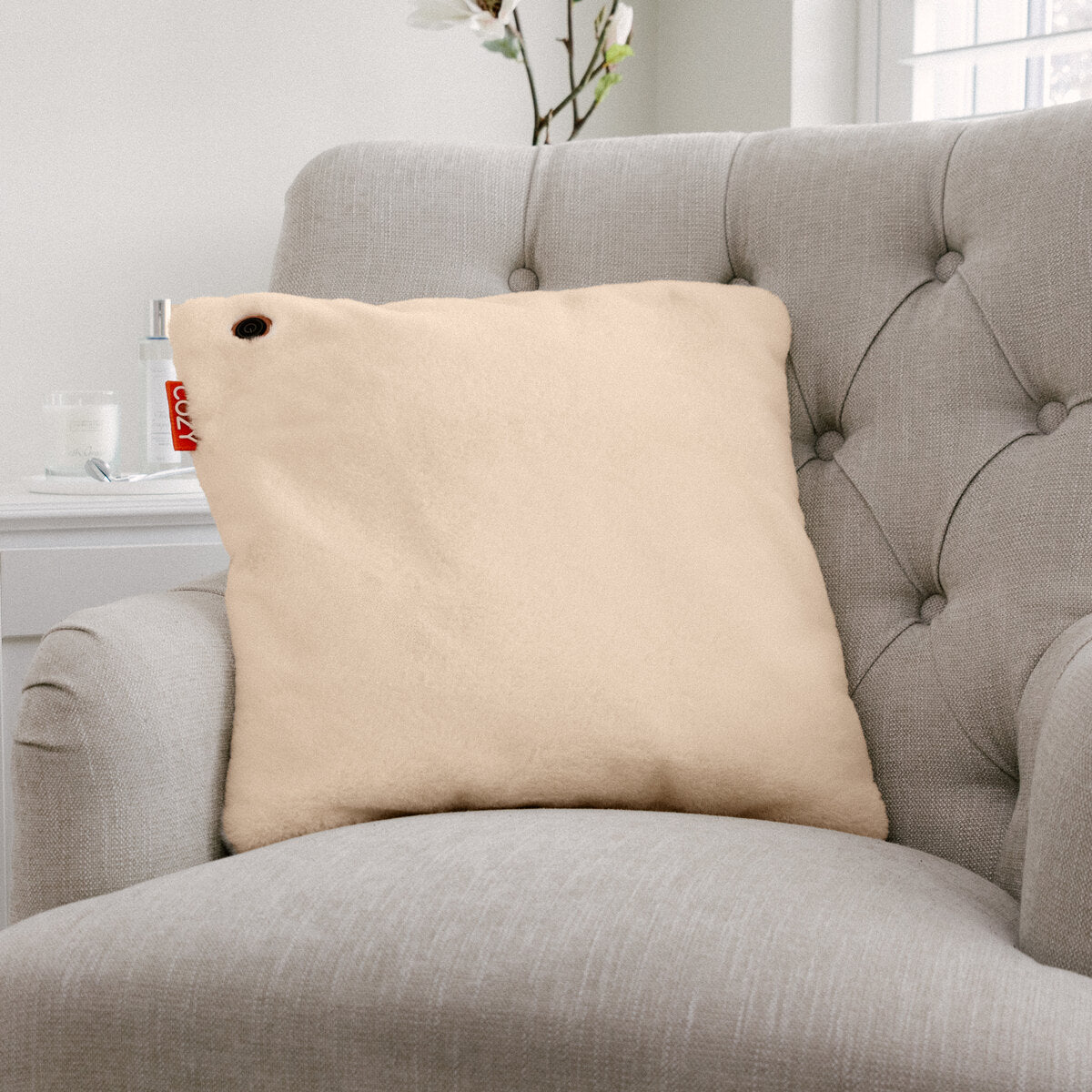 The COZY Heated Cushion Cordless - UNA Faux Fur (45cm x 45cm) by Cozy Cushion EU, a beige square pillow with a red tag and grommet, sits on a light gray tufted armchair in a bright, modern room.