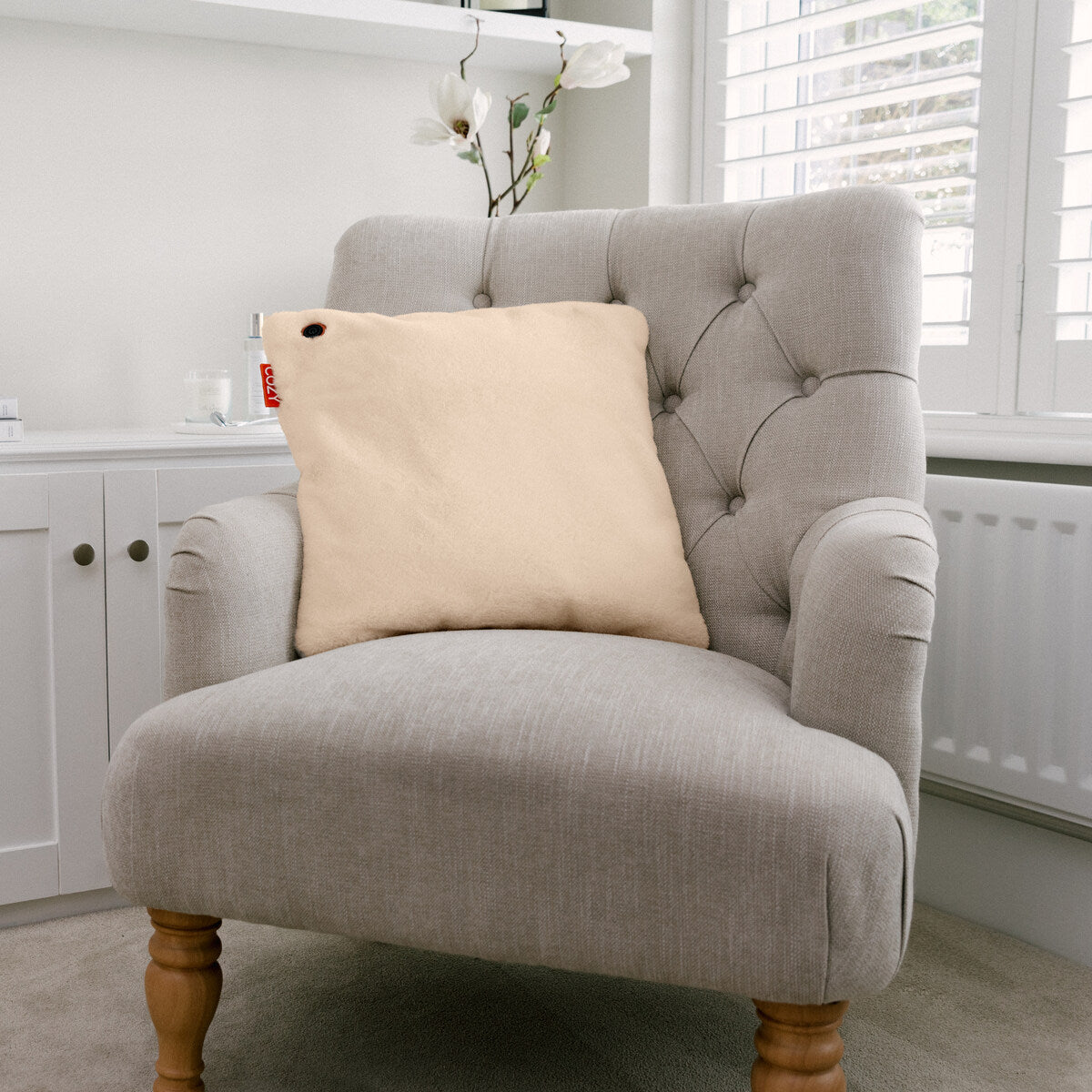 In a bright room, a light grey tufted armchair with wooden legs holds the COZY Heated Cushion Cordless - UNA Faux Fur (45cm x 45cm) by Cozy Cushion EU. White cabinets, flowers, and shuttered windows behind let in natural light.