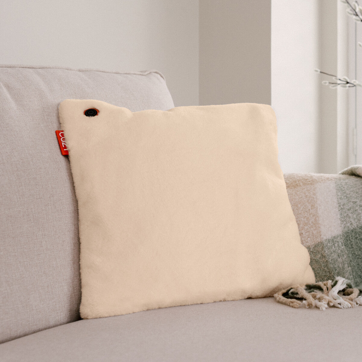 The COZY Heated Cushion Cordless - UNA Faux Fur (45cm x 45cm) by Cozy Cushion EU, cream-colored and square with a red tag and button, sits on a light beige sofa next to a green plaid blanket, with a plant and neutral wall in the background.