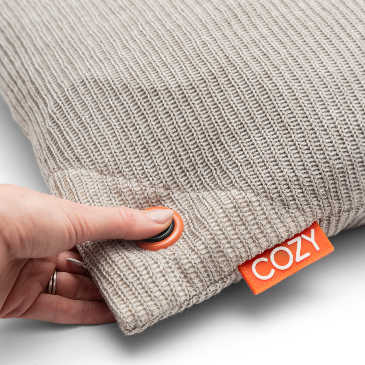 A hand presses a button on the COZY Heated Cushion Cordless - UNA Knitted (45cm x 45cm) by Cozy Cushion EU, highlighting its textured beige knit fabric with an orange COZY tag as the cordless heated cushion is activated for extra comfort.