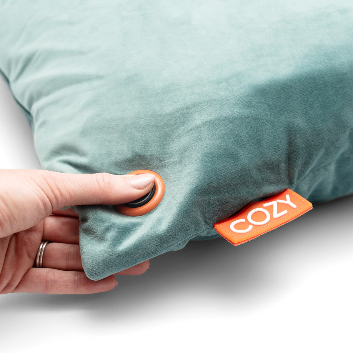 A hand presses a round button on the COZY Heated Cushion Cordless - UNA Velvet (45cm x 45cm) by Cozy Cushion EU, a soft teal-blue cushion featuring an orange COZY tag on its side.