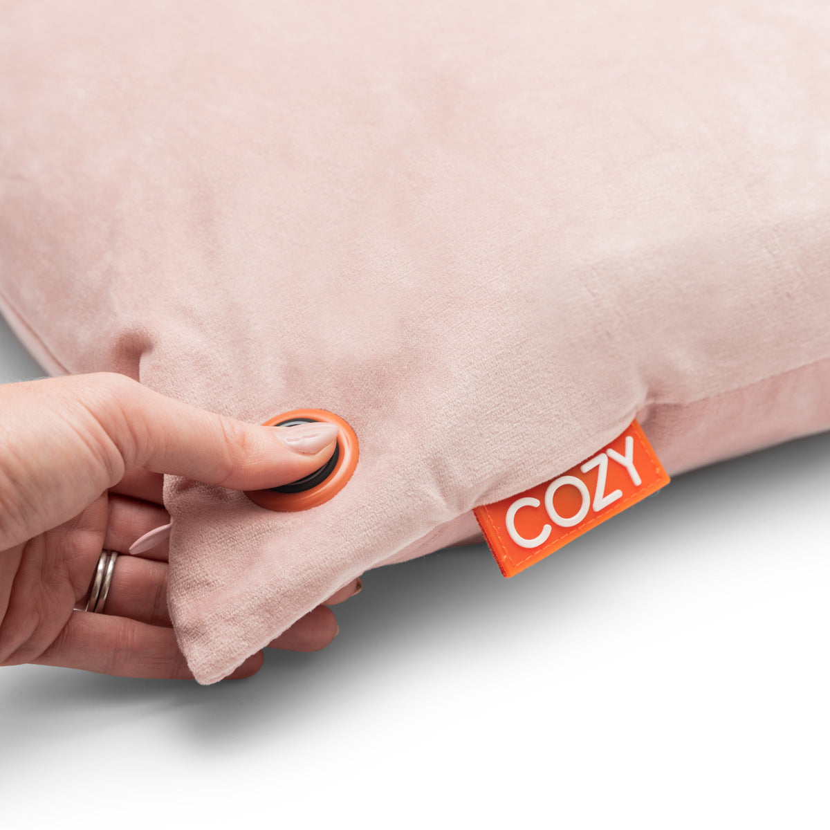 A hand presses a round button on the corner of the Cozy Cushion EU COZY Heated Cushion Cordless - UNA Velvet (45cm x 45cm), a soft, light pink cushion with an orange “COZY” tag.