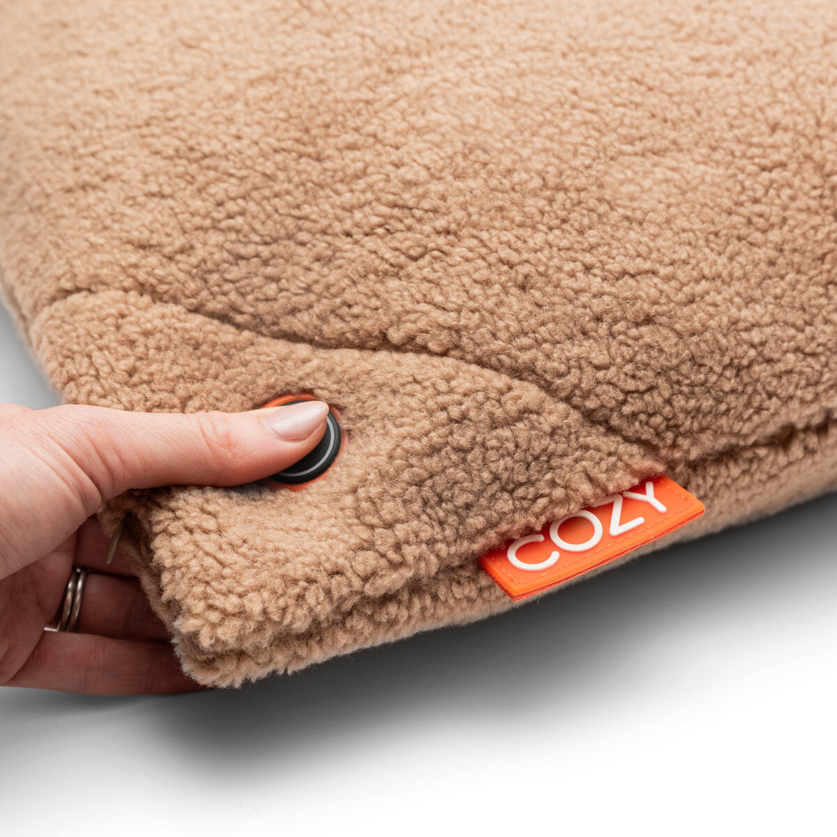 A hand presses a black button on the soft, beige COZY Heated Cushion Cordless - UNA Woolly (45cm x 45cm) by Cozy Cushion EU, activating its cordless warmth.
