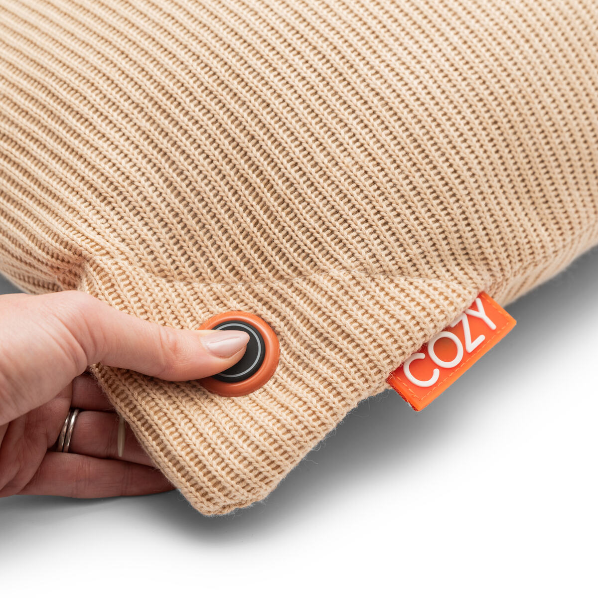 A hand presses a round button on the beige COZY Heated Cushion Cordless - UNA Knitted (45cm x 45cm) by Cozy Cushion EU, showing an orange COZY tag at the seam.