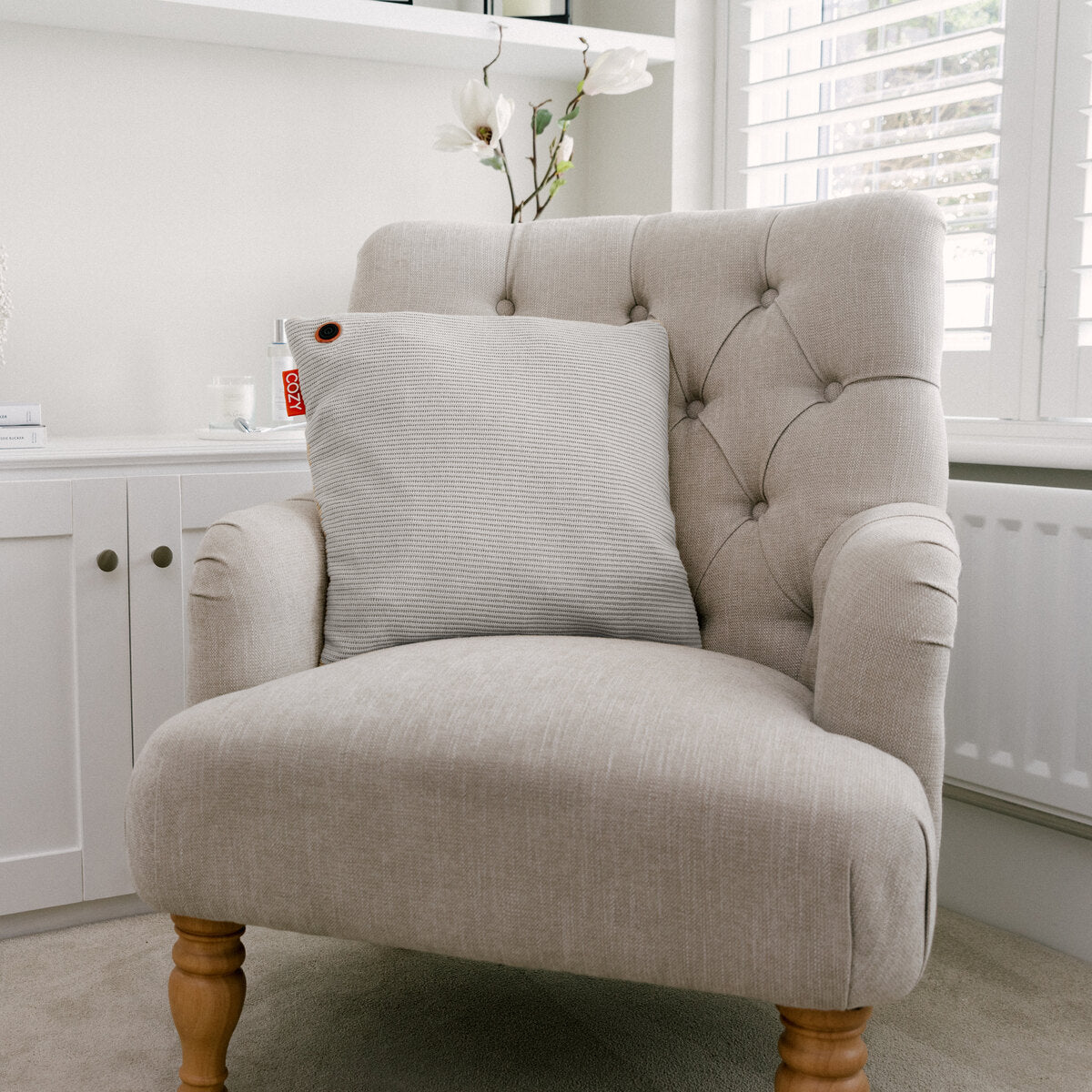 A light beige armchair with wooden legs and a tufted backrest sits in a bright room. A COZY Heated Cushion Cordless - UNA Knitted (45cm x 45cm) by Cozy Cushion EU rests on the chair, with white shelves and a window with shutters behind.