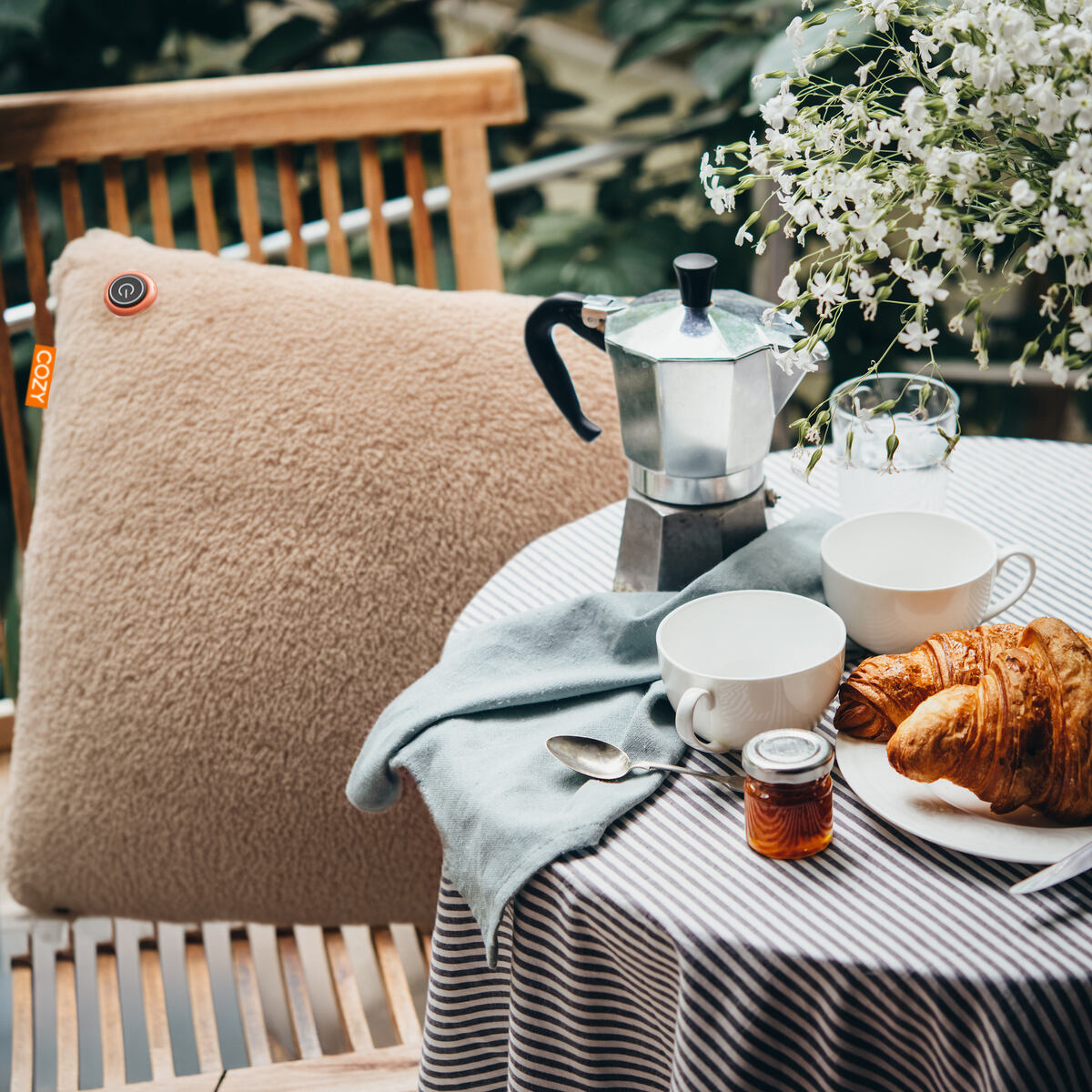 A cozy outdoor table features a striped tablecloth, two white cups, a moka pot, croissants, jam, a glass of water, and the COZY Heated Cushion Cordless - UNA (45cm x 45cm) by Cozy Cushion EU on a wooden chair beside white flowers.